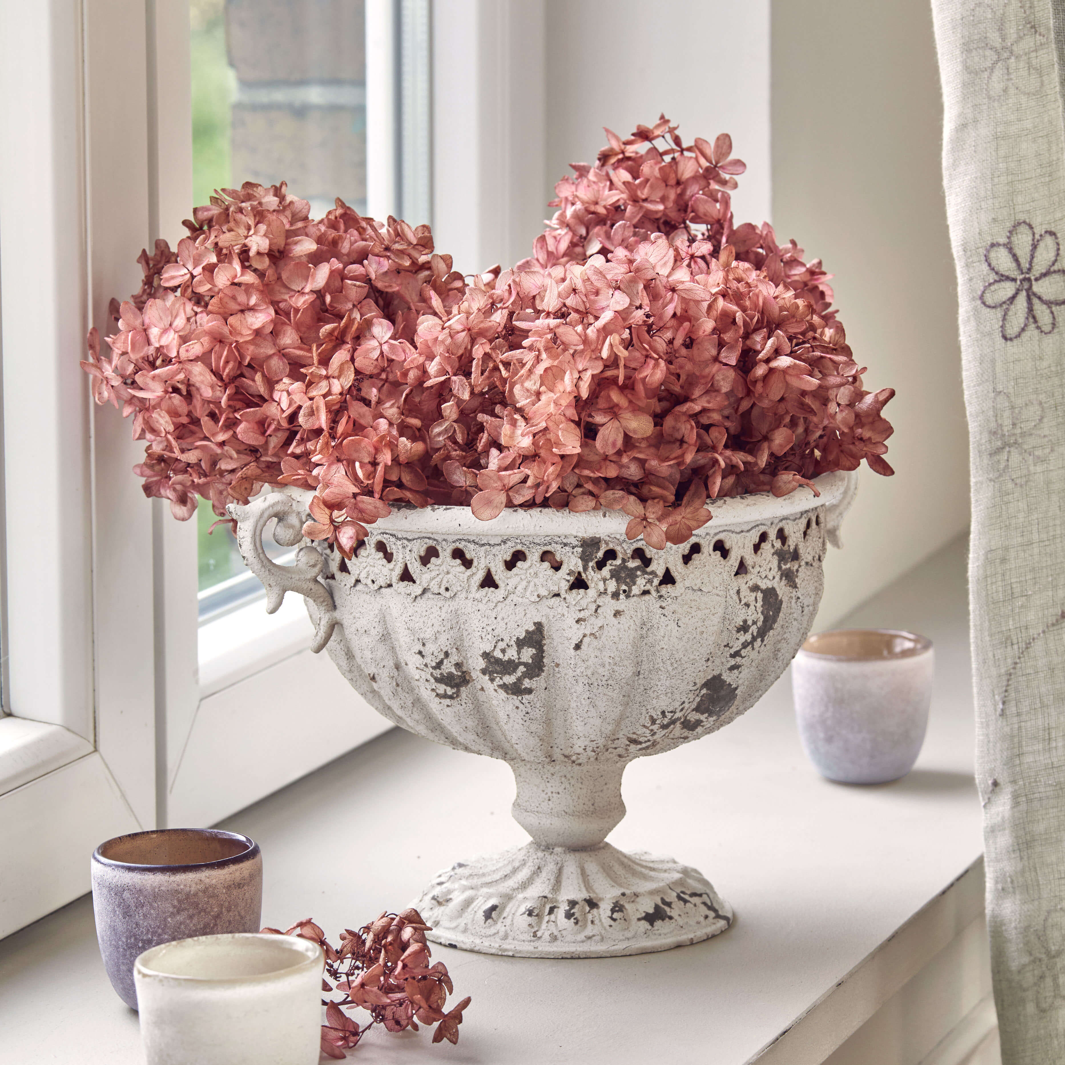 Weiße Metall-Schale mit antikem Finish und getrockneten rosa Blumen auf Fensterbank
