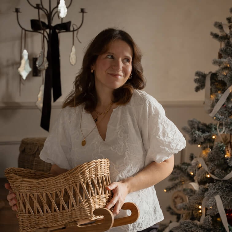 Frau in weißer bestickter Bluse hält einen Korb aus Weide auf Holzschlitten neben geschmücktem Weihnachtsbaum