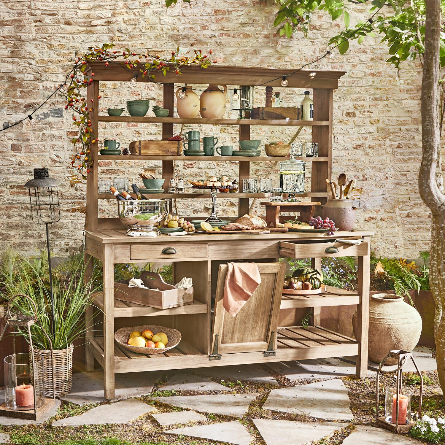 Gartenschrank Fallon Holz-Gartenmöbel mit Regalen und Schubladen, dekoriert mit Geschirr und Speisen auf Terrasse