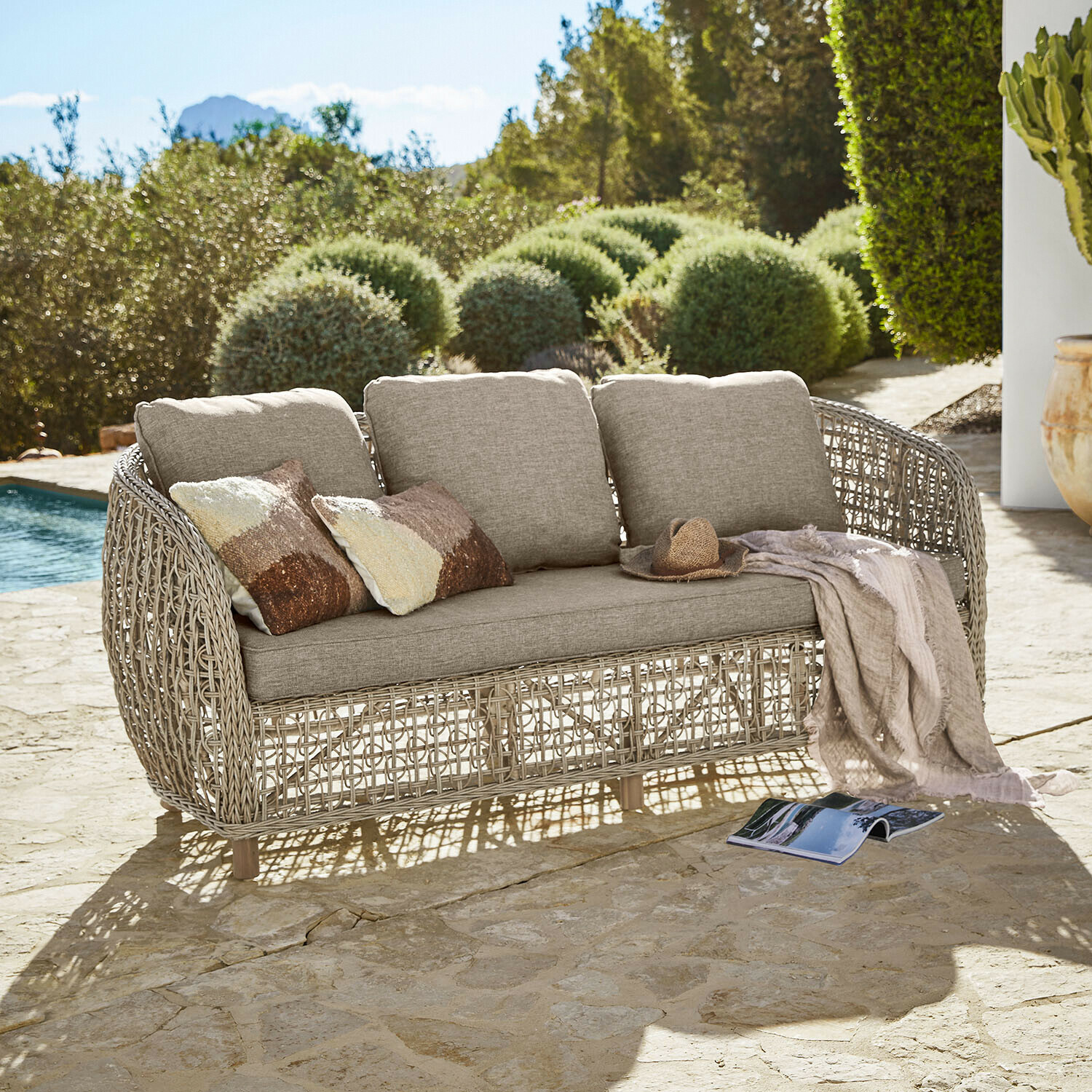 Gartensofa mit beigen Polstern und geflochtenem Rattangestell auf steinerner Terrasse am Pool