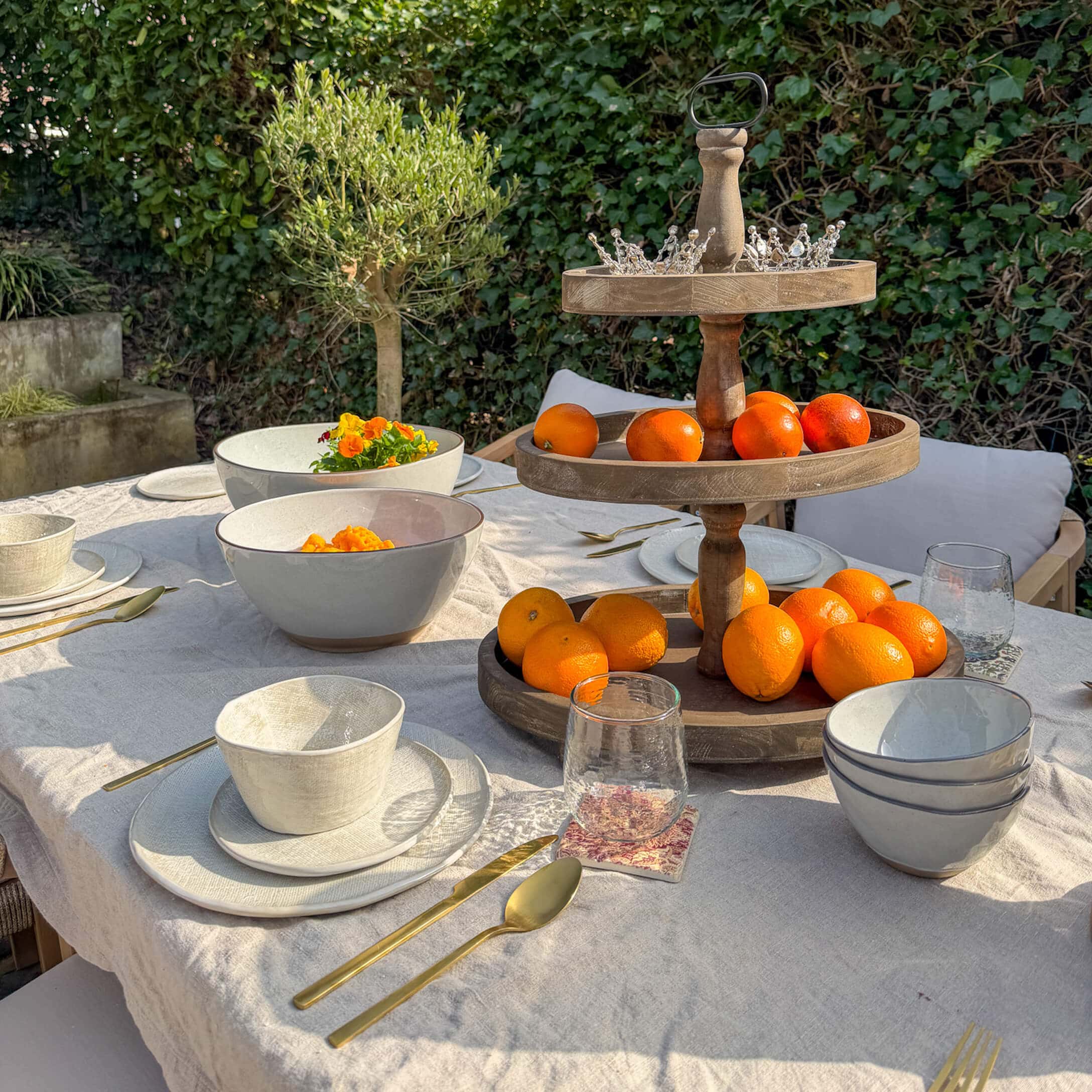 Gartentisch mit weißem Keramikgeschirr, goldenen Bestecken und hölzernem Obstständer mit Orangen