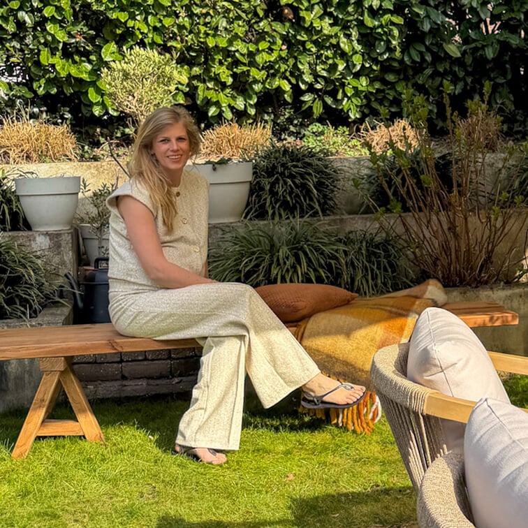 Frau in hellem Outfit sitzt auf einer Holzbank im Garten mit grünen Pflanzen und Kissen