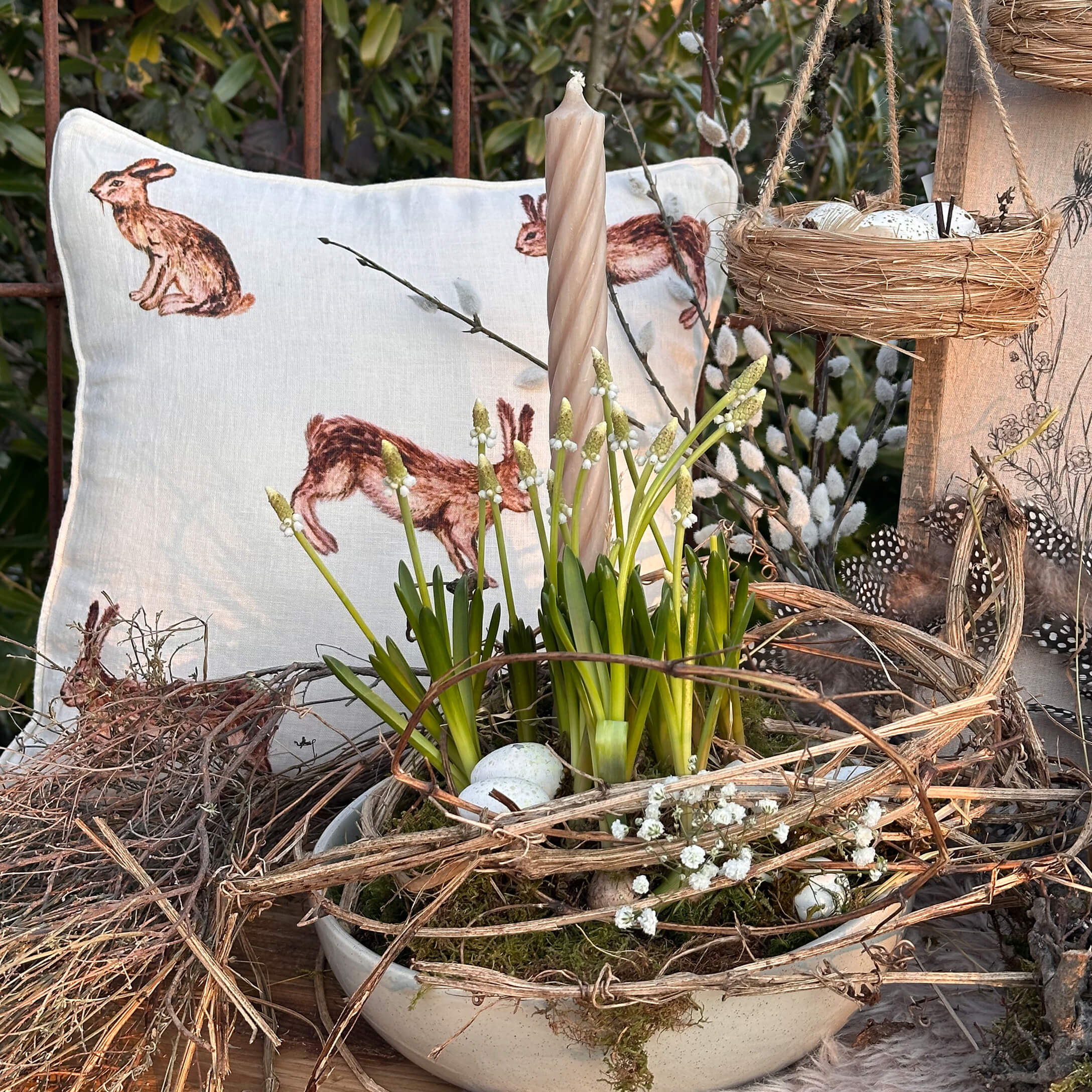 Natürlich dekorierte Osterfreude mit weißer Kerze, grünen Blumen und Hasenmotiv-Kissen auf Holztisch