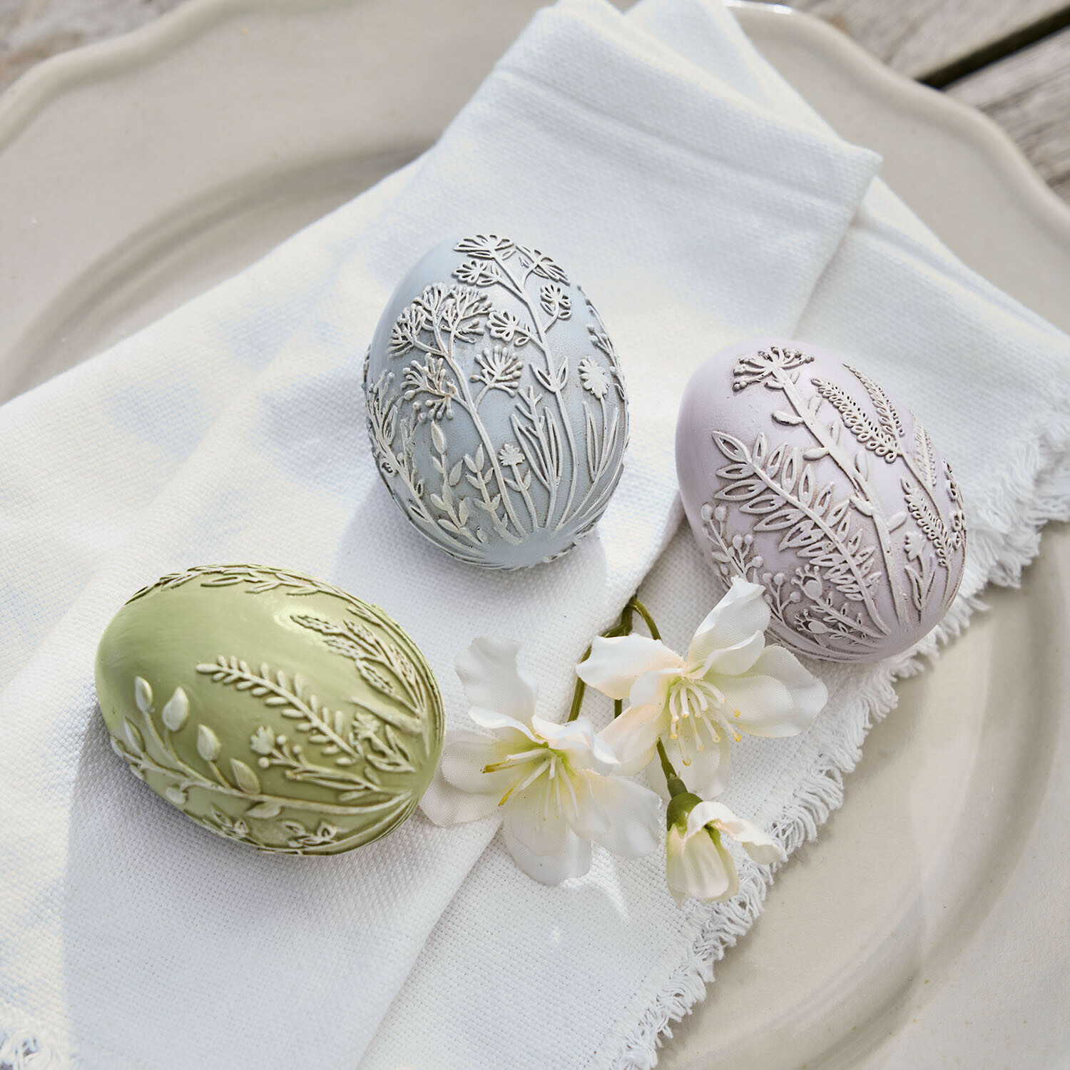 Drei pastellfarbene Dekoeier mit pflanzlichem Relief auf weißem Stoff und Blumen auf Teller