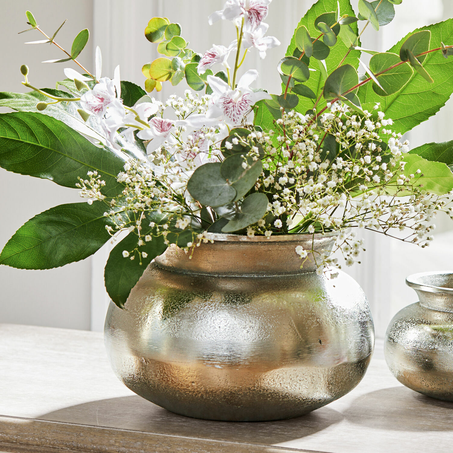 Vase Hadrien Silberne Metallvase mit weißen Blumen und grünen Blättern auf Holztisch, Wohnaccessoire