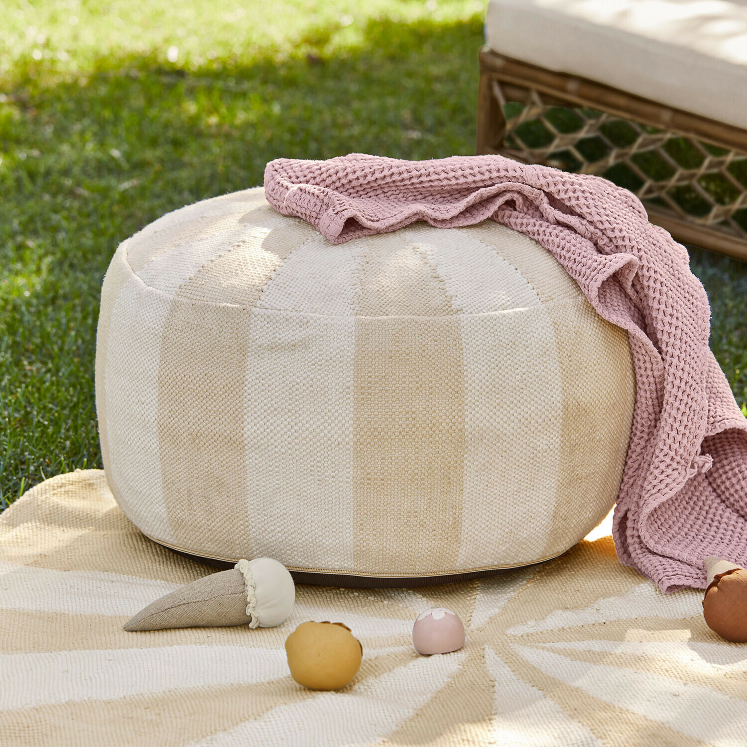 Beiger runder Stoff-Pouf mit rosa Strickdecke und Spielzeug auf Gras im Garten
