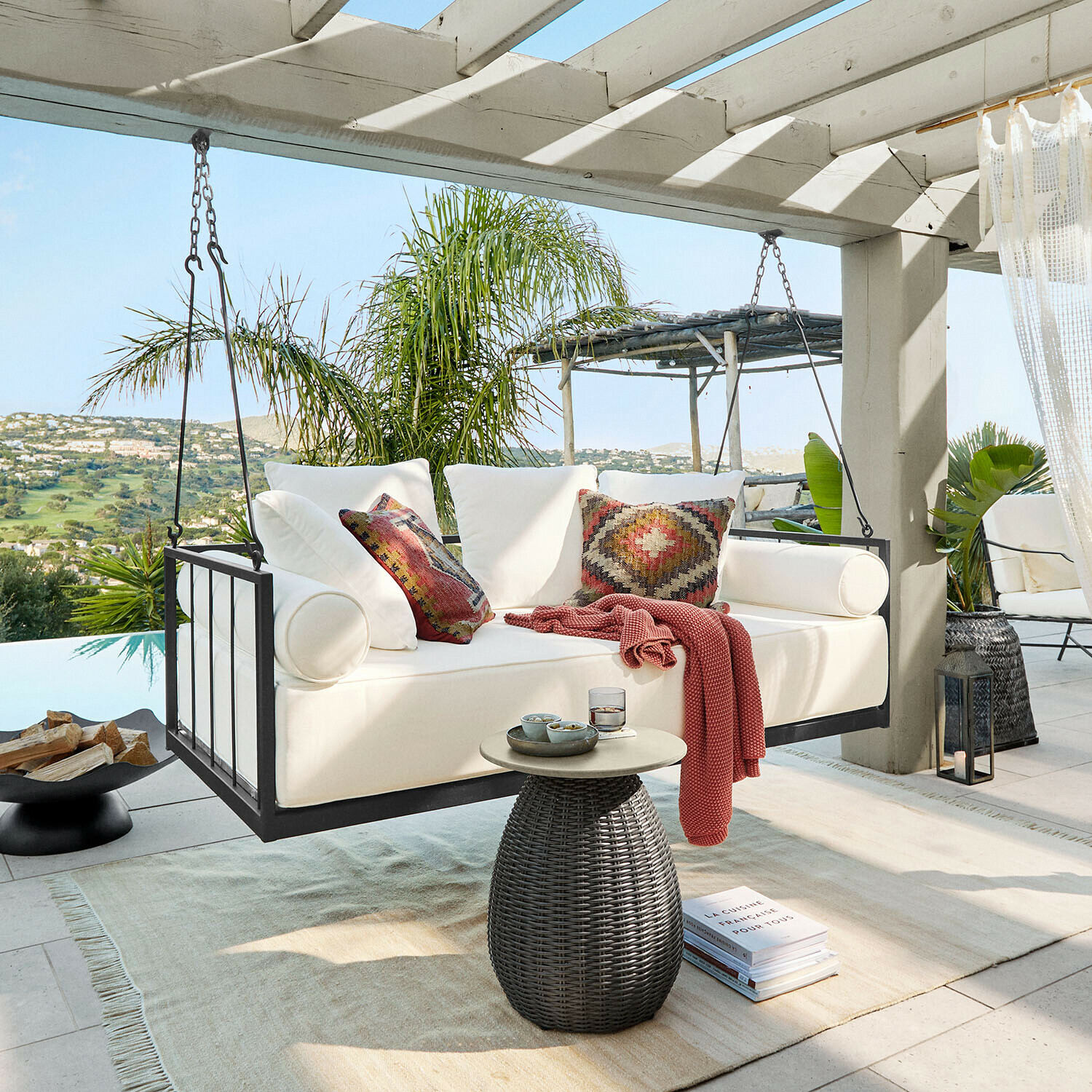 Hängebank Bellecôtes Weiße Hängesofa mit Metallrahmen und bunten Kissen auf Terrasse mit grünem Ausblick
