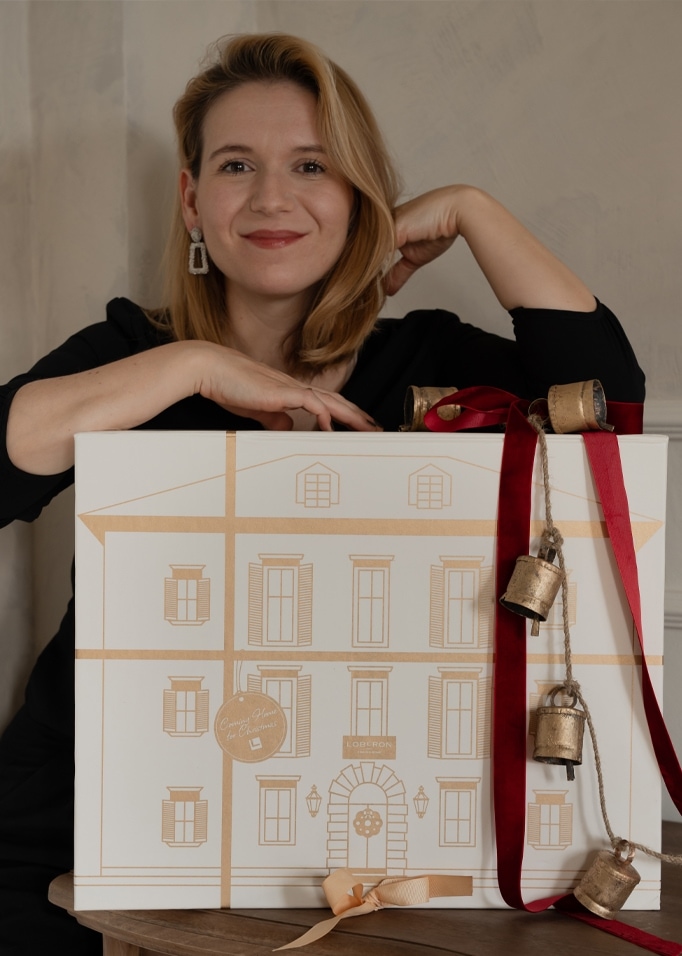 Frau mit Geschenkbox in Weiß mit goldenem Hausdruck und roten Glocken an Schnur als Adventsdeko