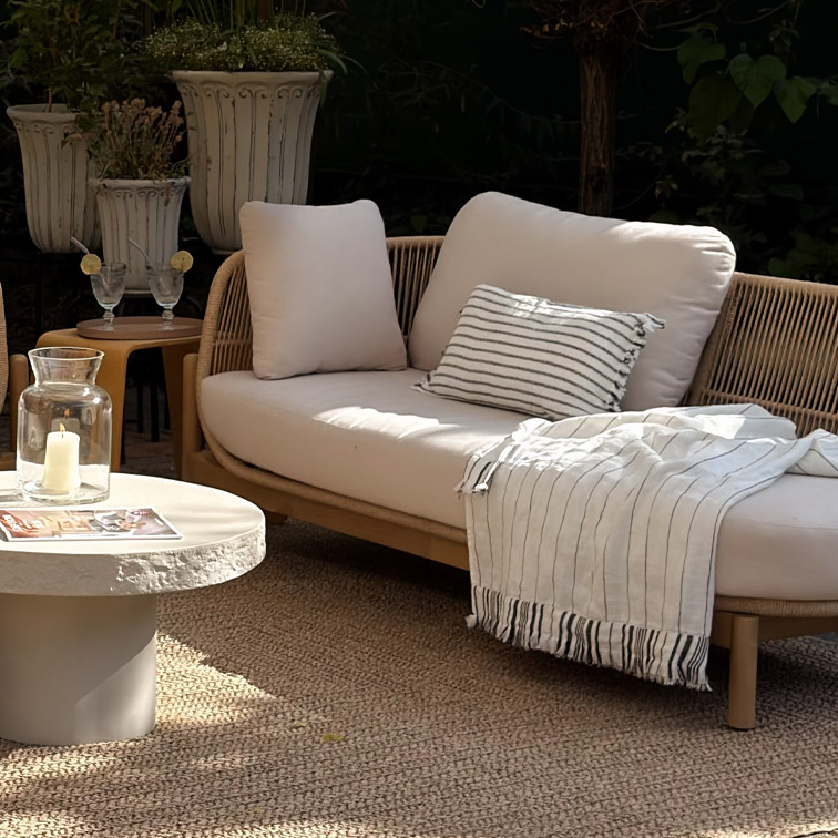 Moderne Outdoor-Sofa mit hellen Kissen und gestreiftem Plaid auf natürlichem Teppich im Garten