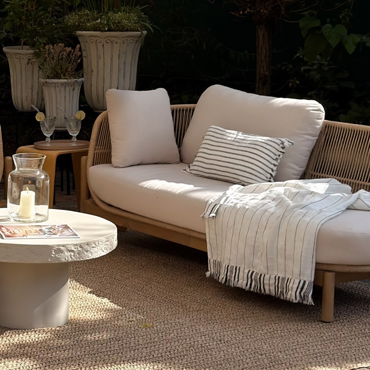 Moderne Outdoor-Sofa mit hellen Kissen und gestreiftem Plaid auf natürlichem Teppich im Garten