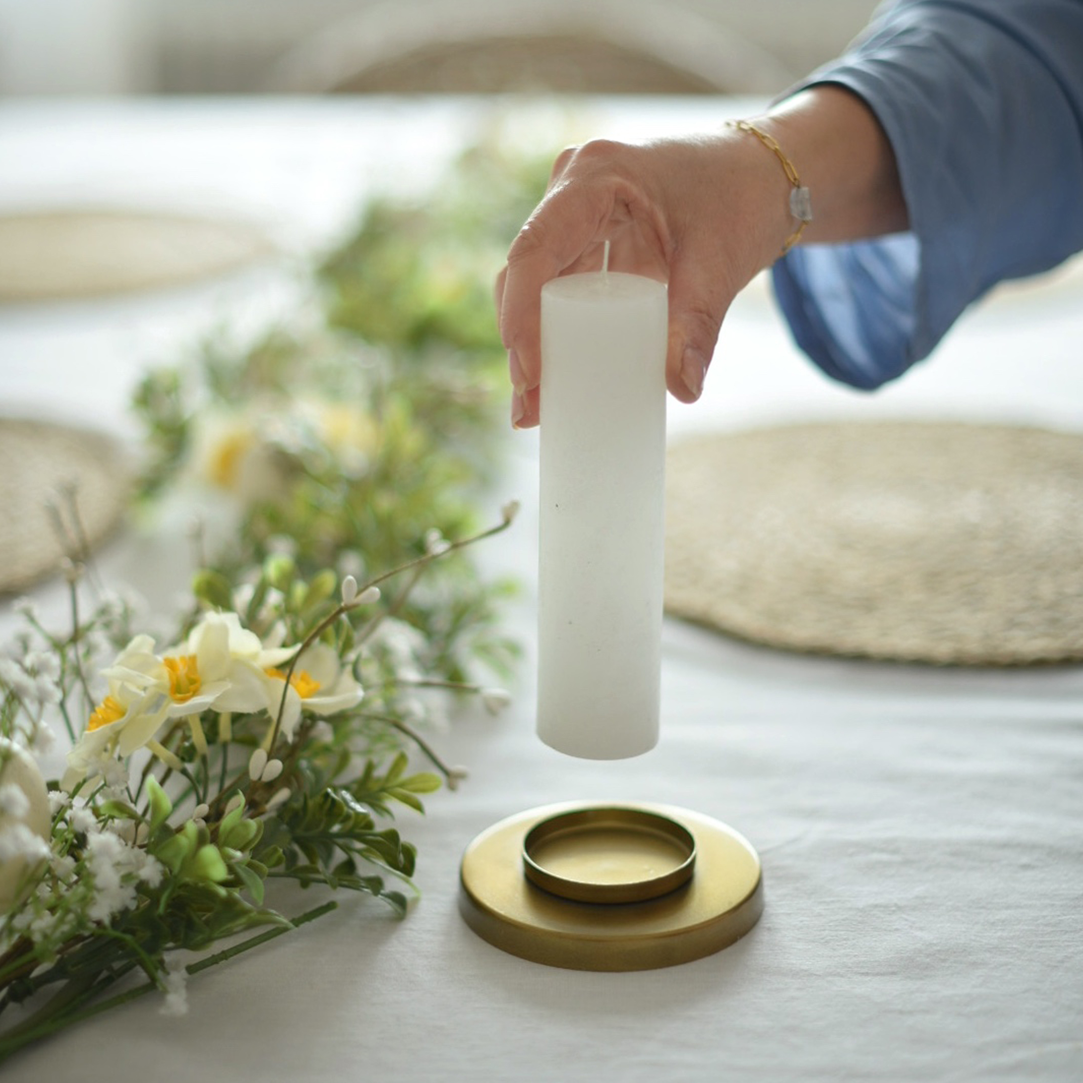 Weiße Stumpenkerze über goldenem Kerzenhalter mit Blumenarrangement auf weißem Tischdecke