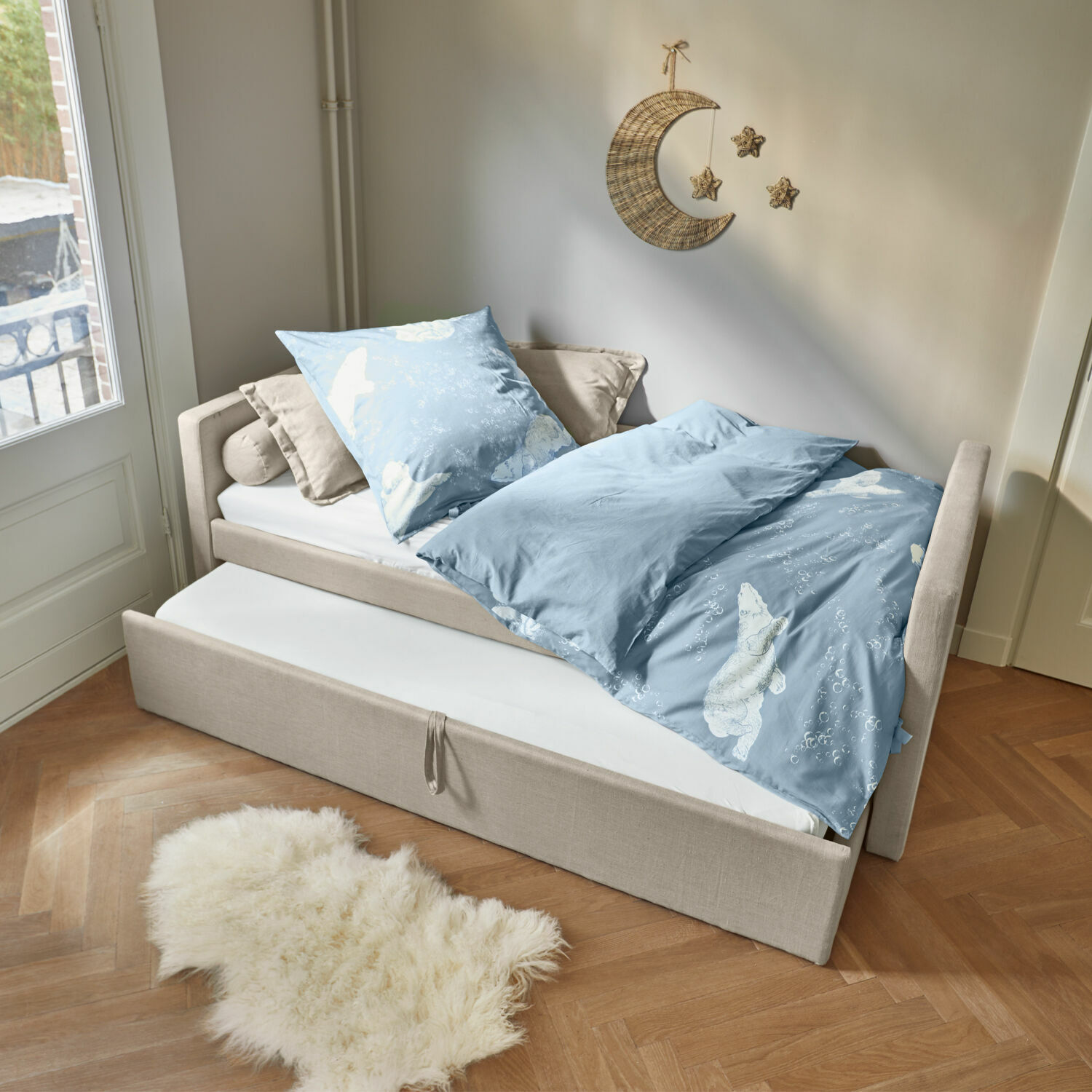 Bett Thistlewood Beiges Bett mit ausziehbarer Matratze und blauer Bettwäsche mit Eisbärmotiv in hellem Zimmer.