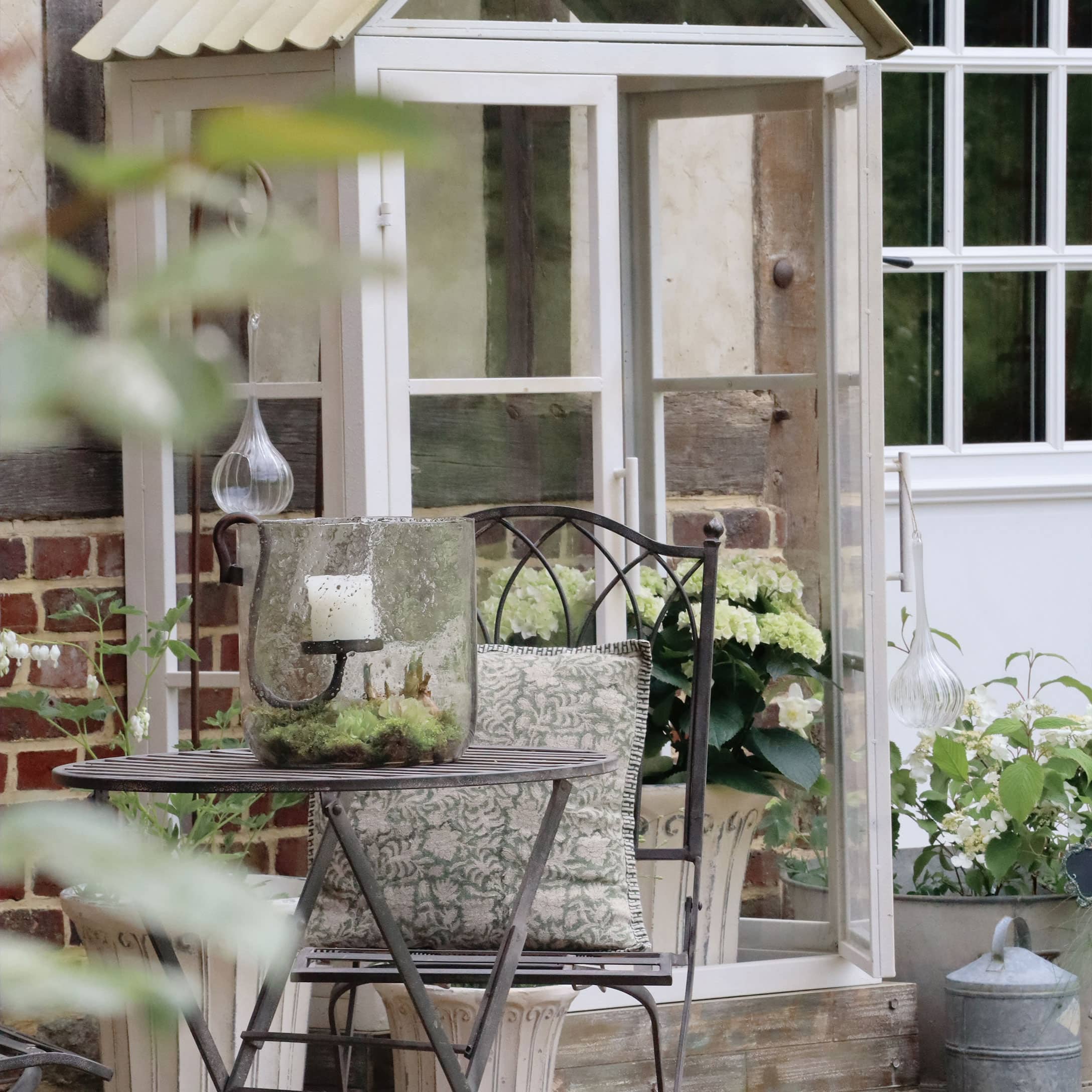 Garten mit Metalltisch und Stuhl mit Kissen, Glaslaterne mit weißer Kerze und weißen Blumen