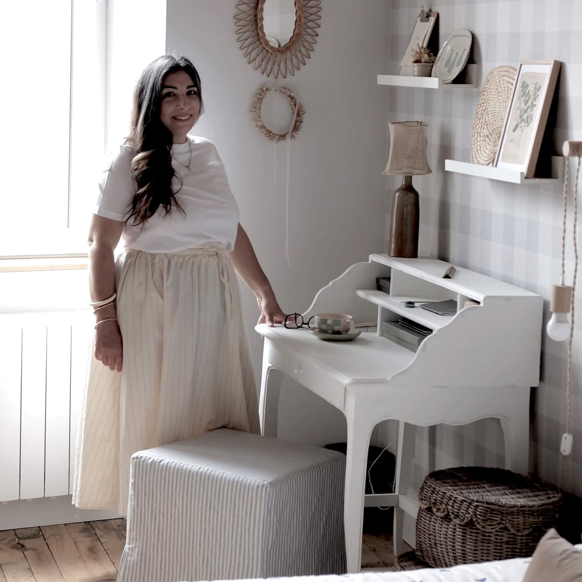 Frau in hellem Rock steht neben weißem Holzschreibtisch in gemütlichem Raum mit natürlichen Dekorationen