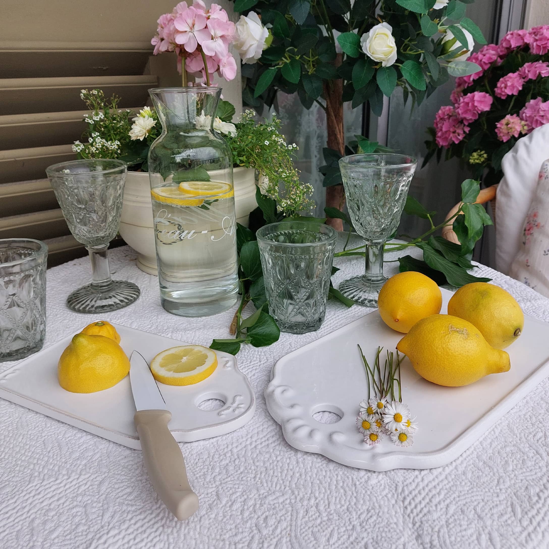 Glaskaraffe mit Zitronenwasser auf weißer Tischdecke, dekorative Gläser und weiße Keramik-Schneidebretter