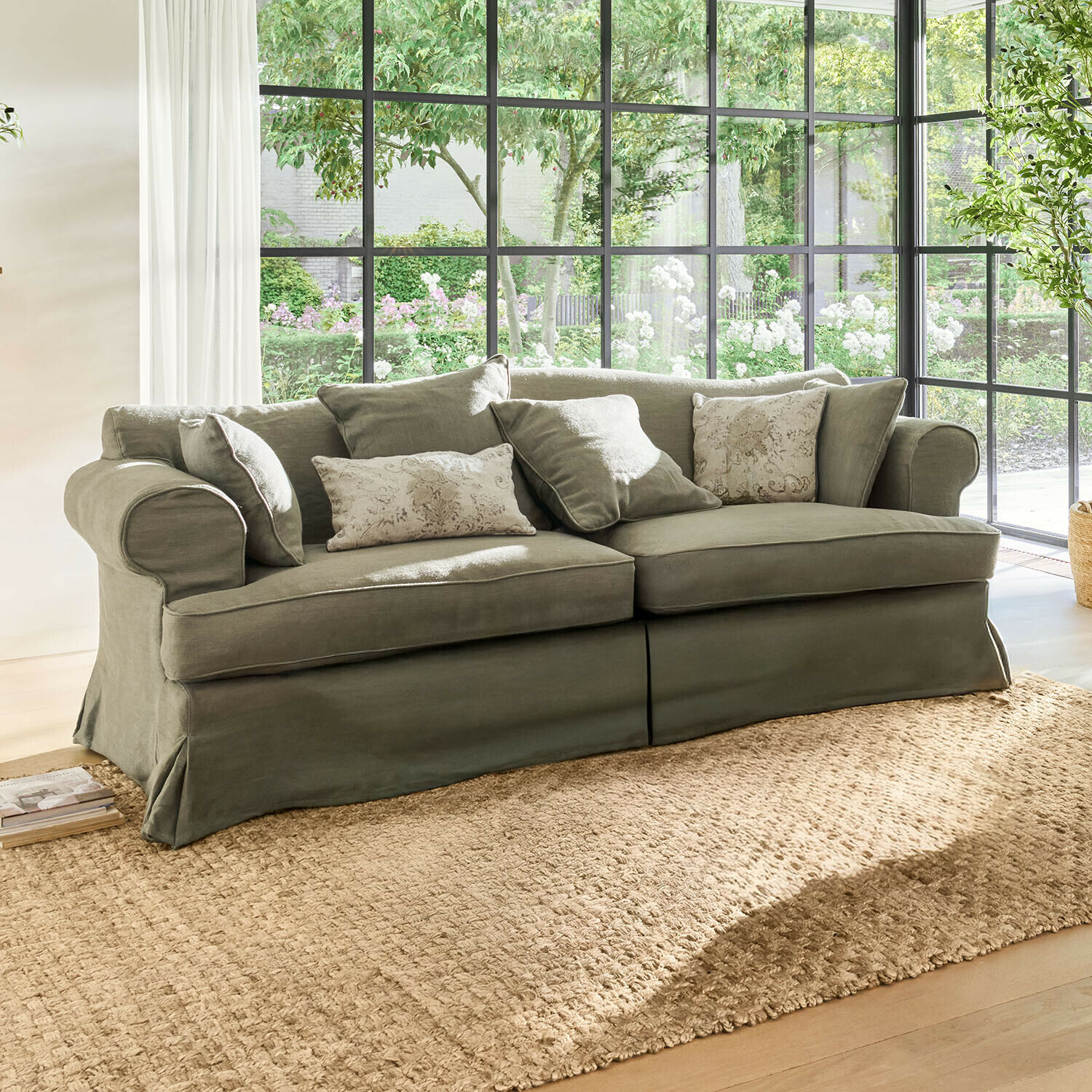 Graues Sofa aus Stoff mit dekorativen Kissen vor großen Fenstern mit Tageslicht