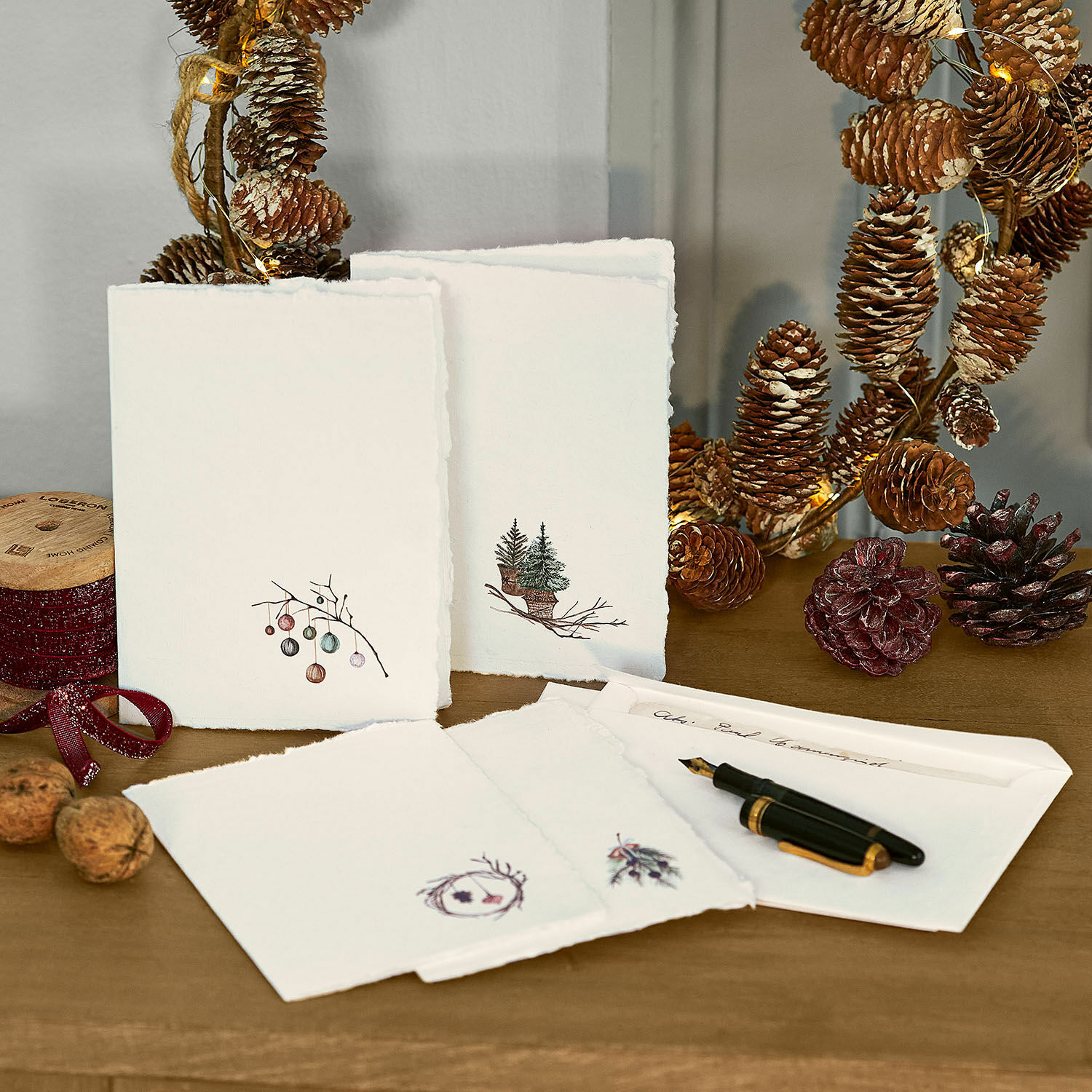 Grußkarte 4er Set Velorine Set Weihnachtskarten aus Papier mit Tannen- und Zapfenmotiven auf Holztisch mit Füllfederhalter und Dekoration