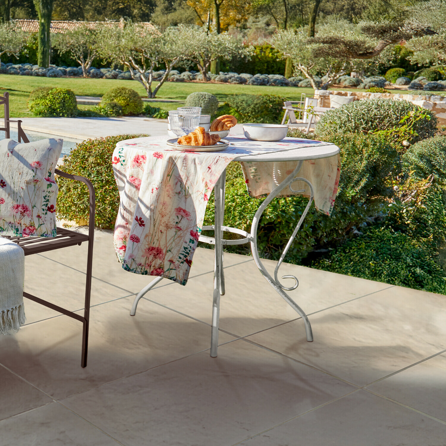 Runder weißer Metall-Gartentisch mit floralem Tischtuch und Gebäck auf Terrasse mit Gartenblick