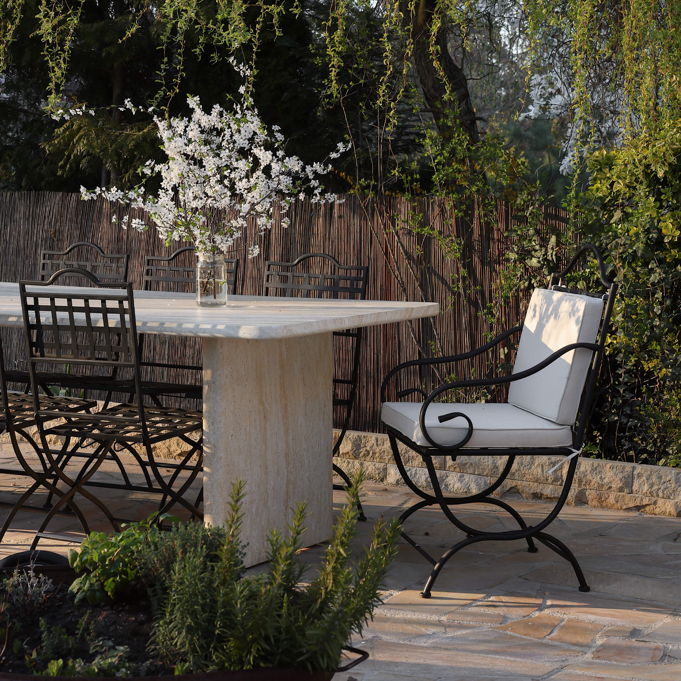 Gartentisch aus Stein mit schwarzen Metallstühlen und hellen Polstern, Blumendekor