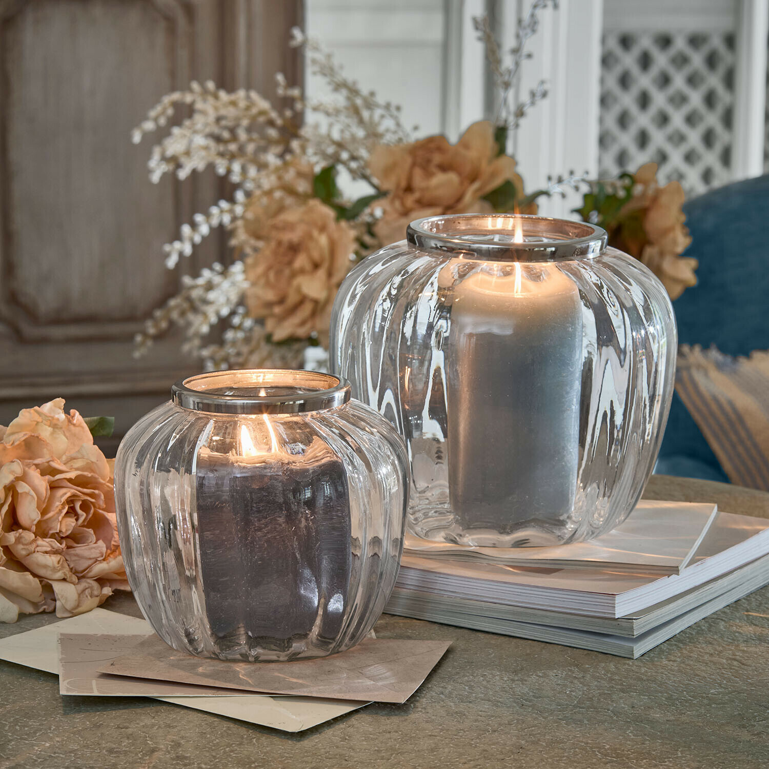 Zwei transparente Glaswindlichter mit Metallrand und grauen Kerzen auf dekorativem Tisch