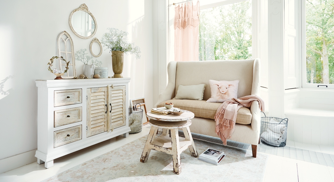 Gemütliches Wohnzimmer im Shabby-Chic mit weißem Holzsideboard und beigem Sofa mit Kissen und rosa Decke