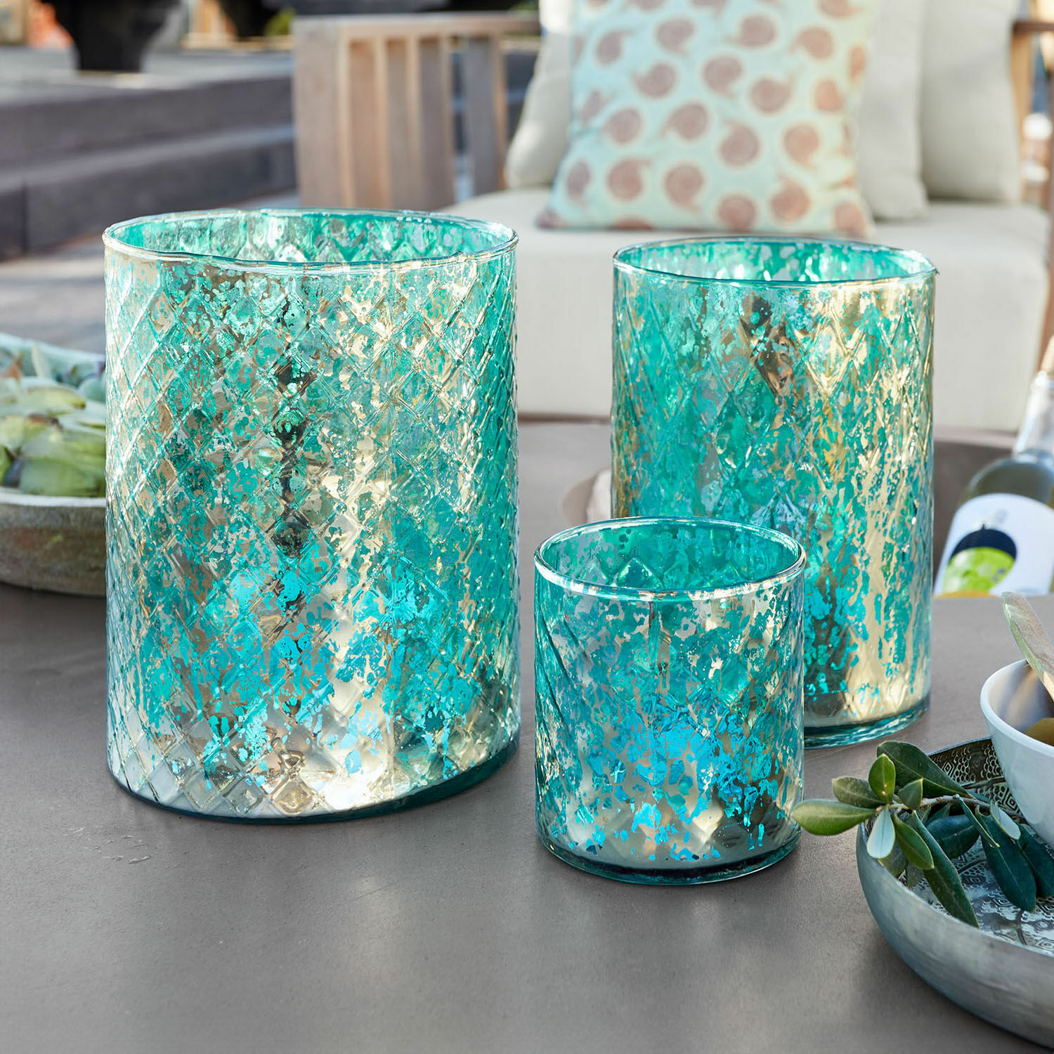 Windlicht 3er Set Firalen Set aus drei türkisfarbenen Glaswindlichtern mit metallischem Muster auf Gartentisch