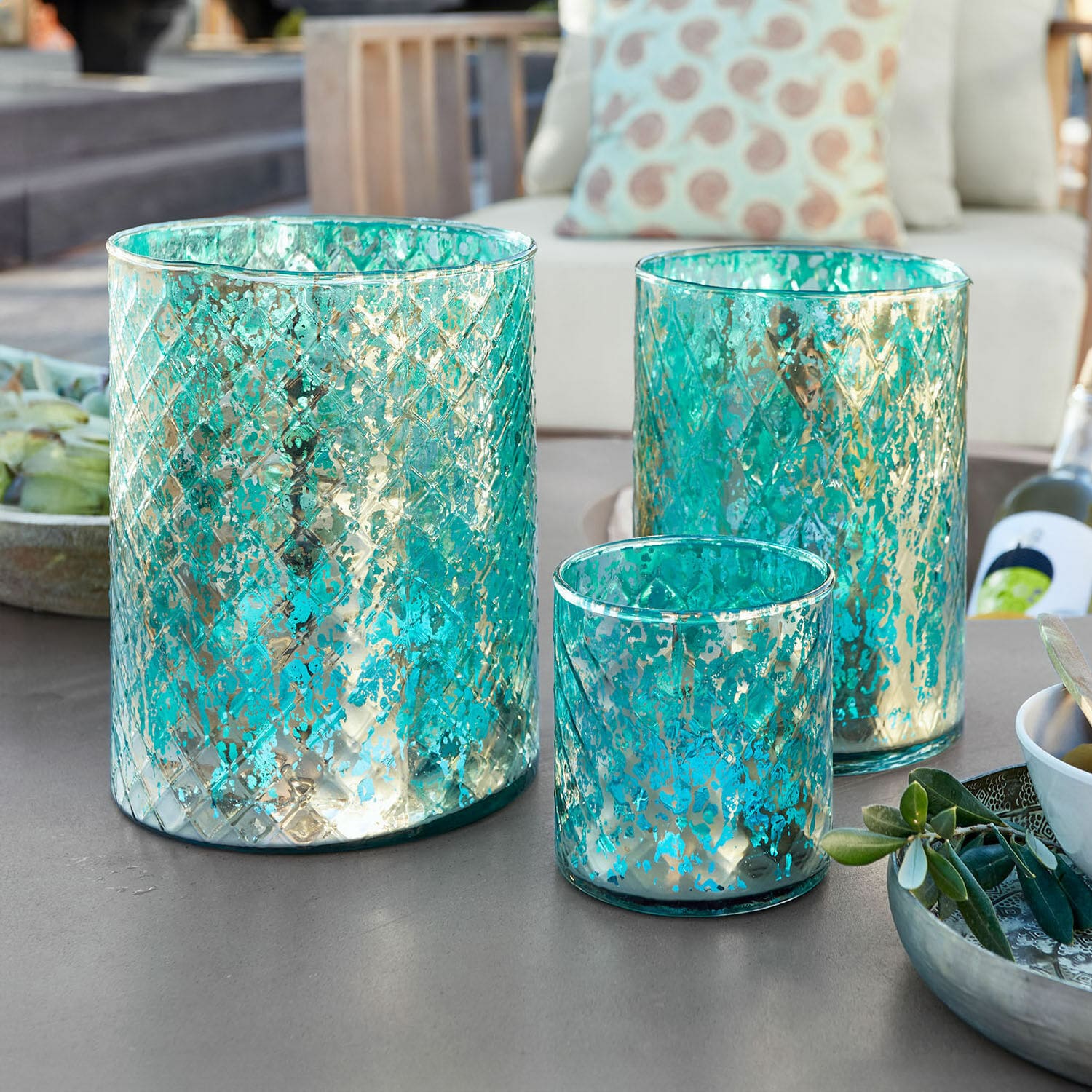 Windlicht 3er Set Firalen Set aus drei türkisfarbenen Glaswindlichtern mit metallischem Muster auf Gartentisch