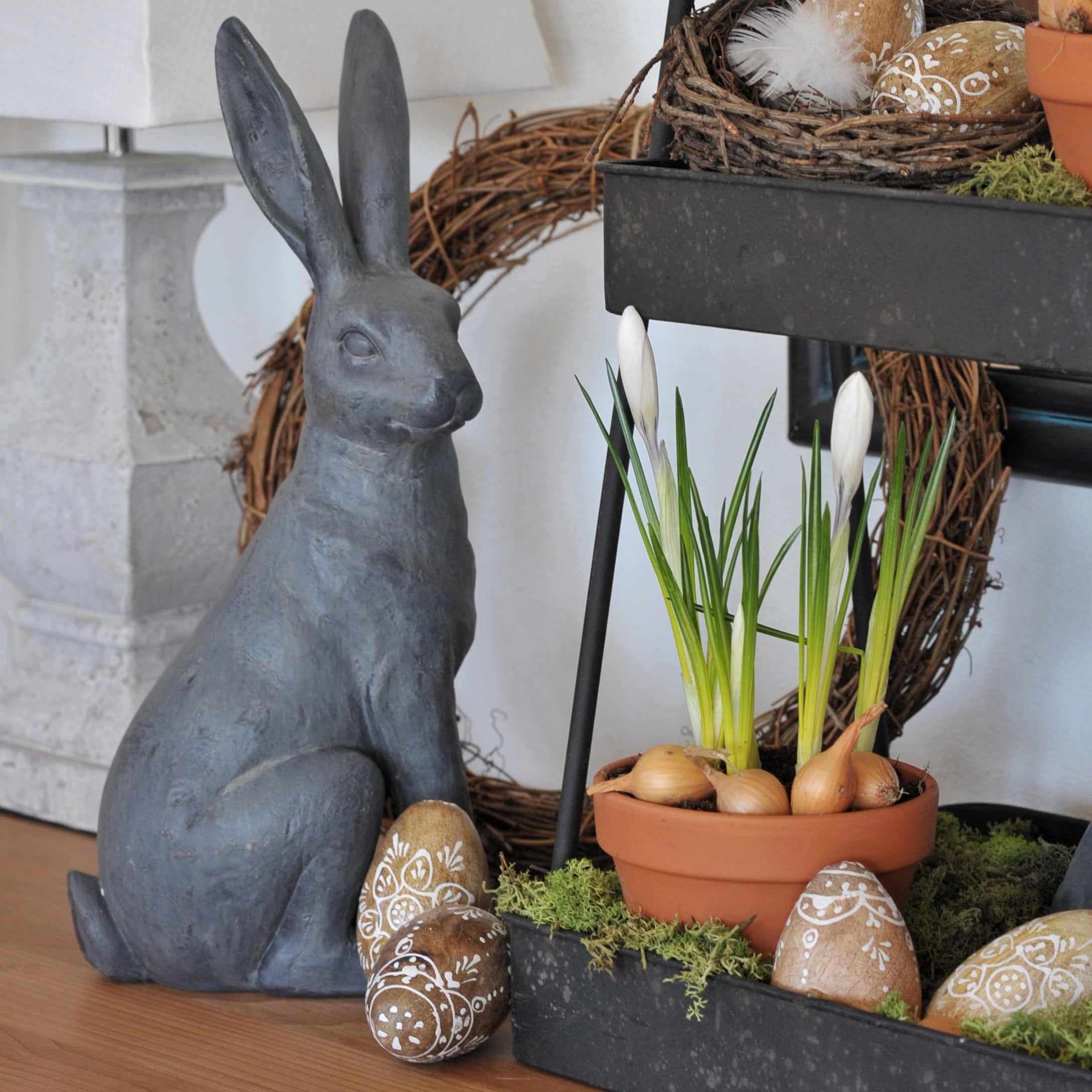 Graue Keramik-Hasenfigur neben Blumentopf mit weißen Blüten und dekorativen Ostereiern für Frühlingsdeko