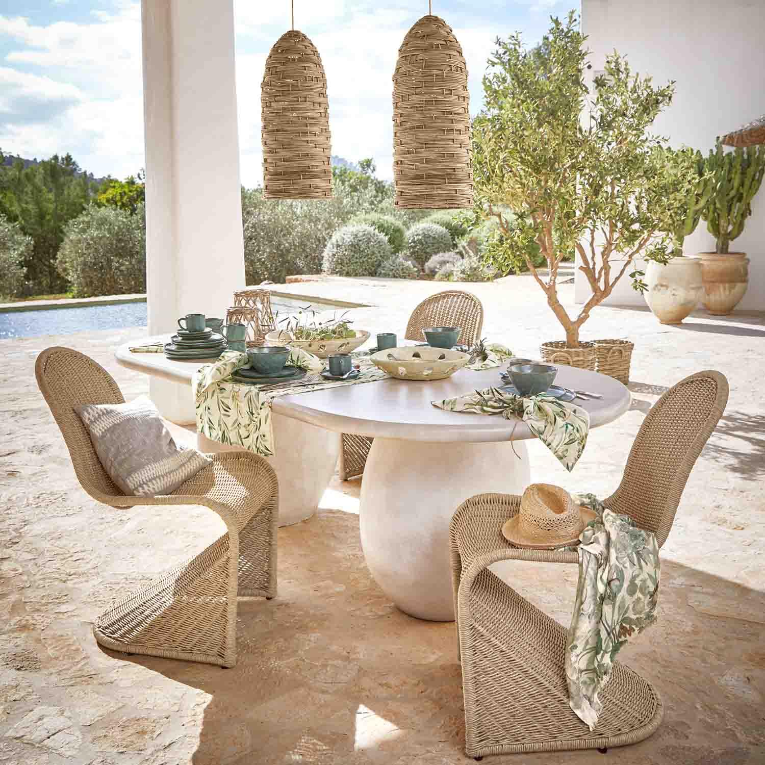 Ovaler weißer Steintisch mit Rattanstühlen und natürlichen Hängeleuchten in sonnigem Garten