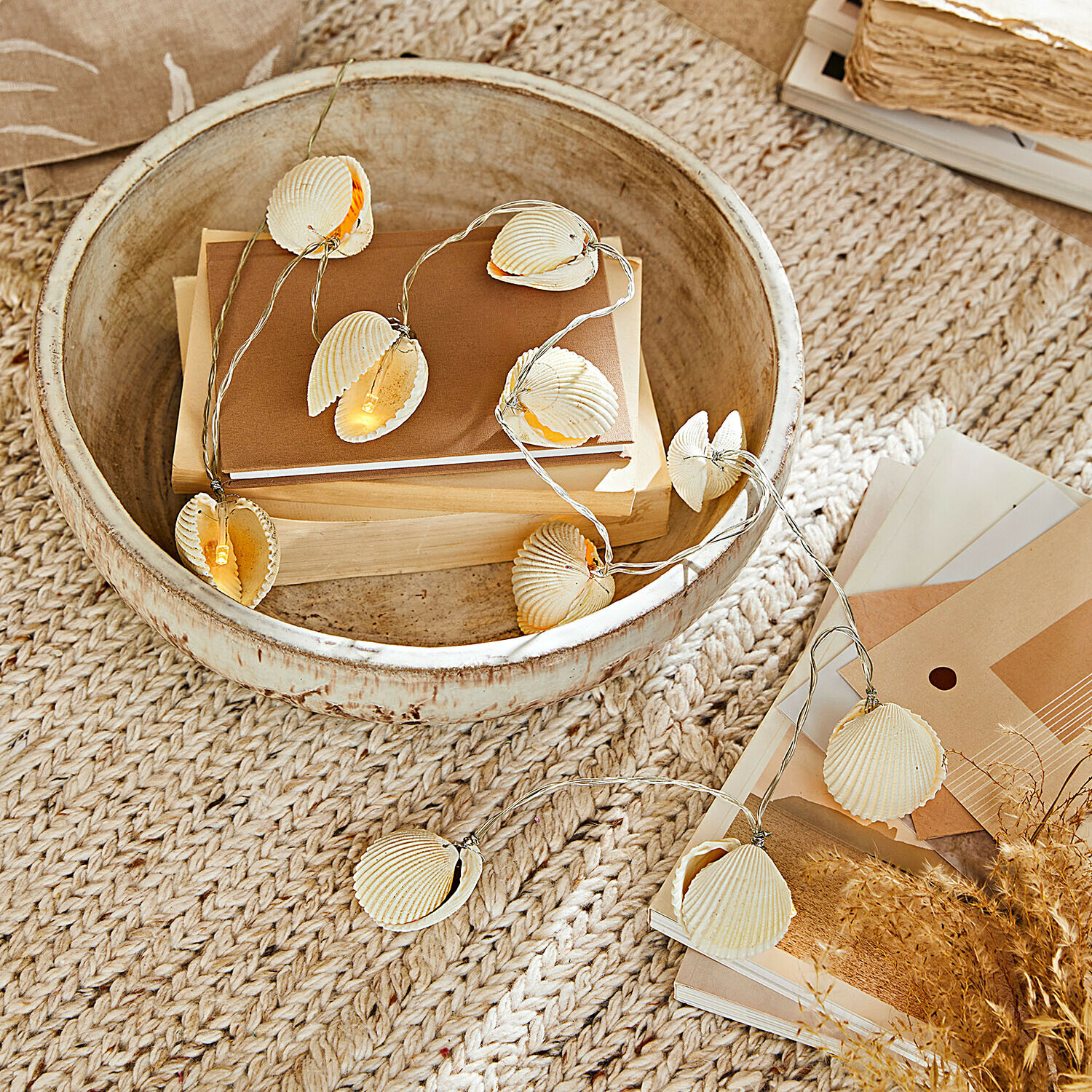 Lichterkette mit natürlichen Muscheln auf beigem Teppich mit Holzschale und Büchern dekorativ drapiert