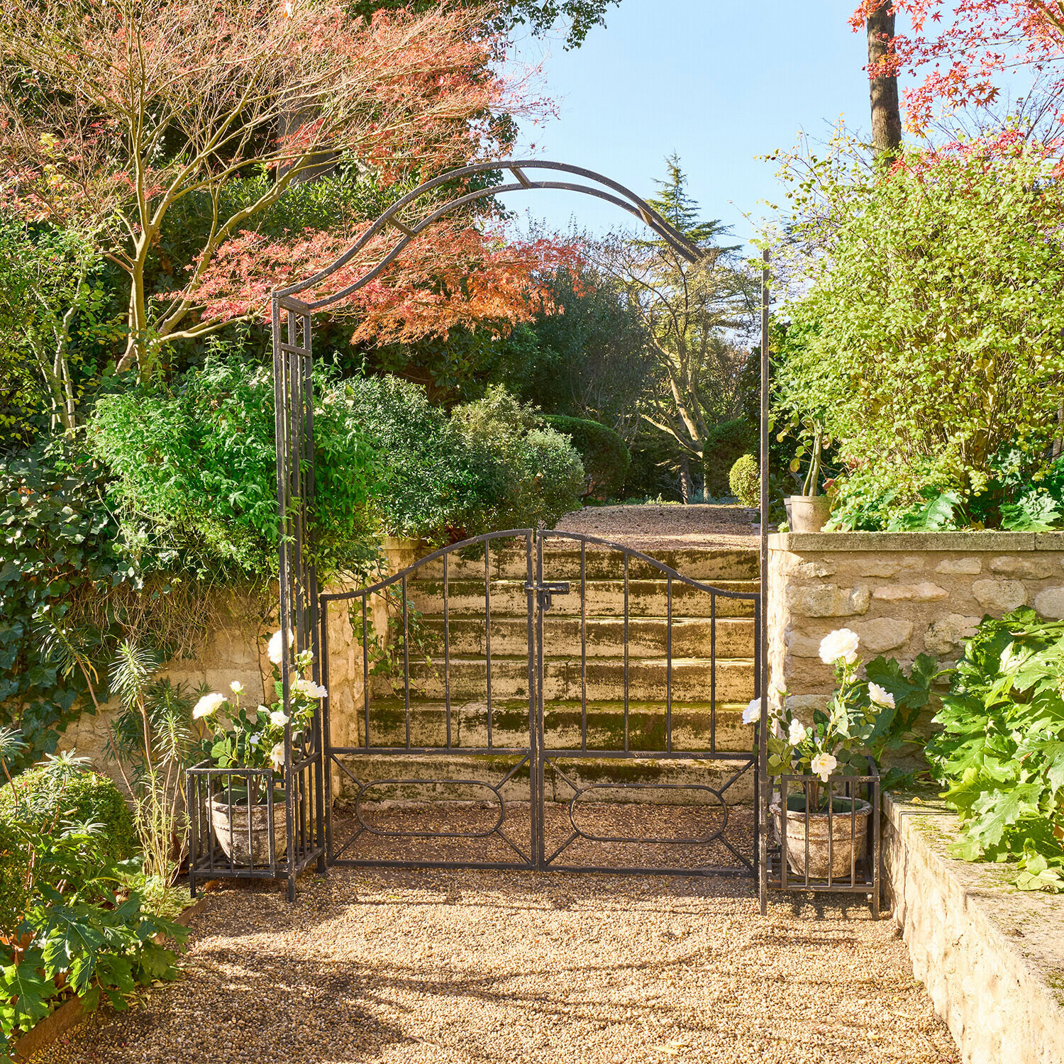 Schwarzes Metall-Gartentor mit Bogen und weißen Blumentöpfen auf sonnigem Kiesweg