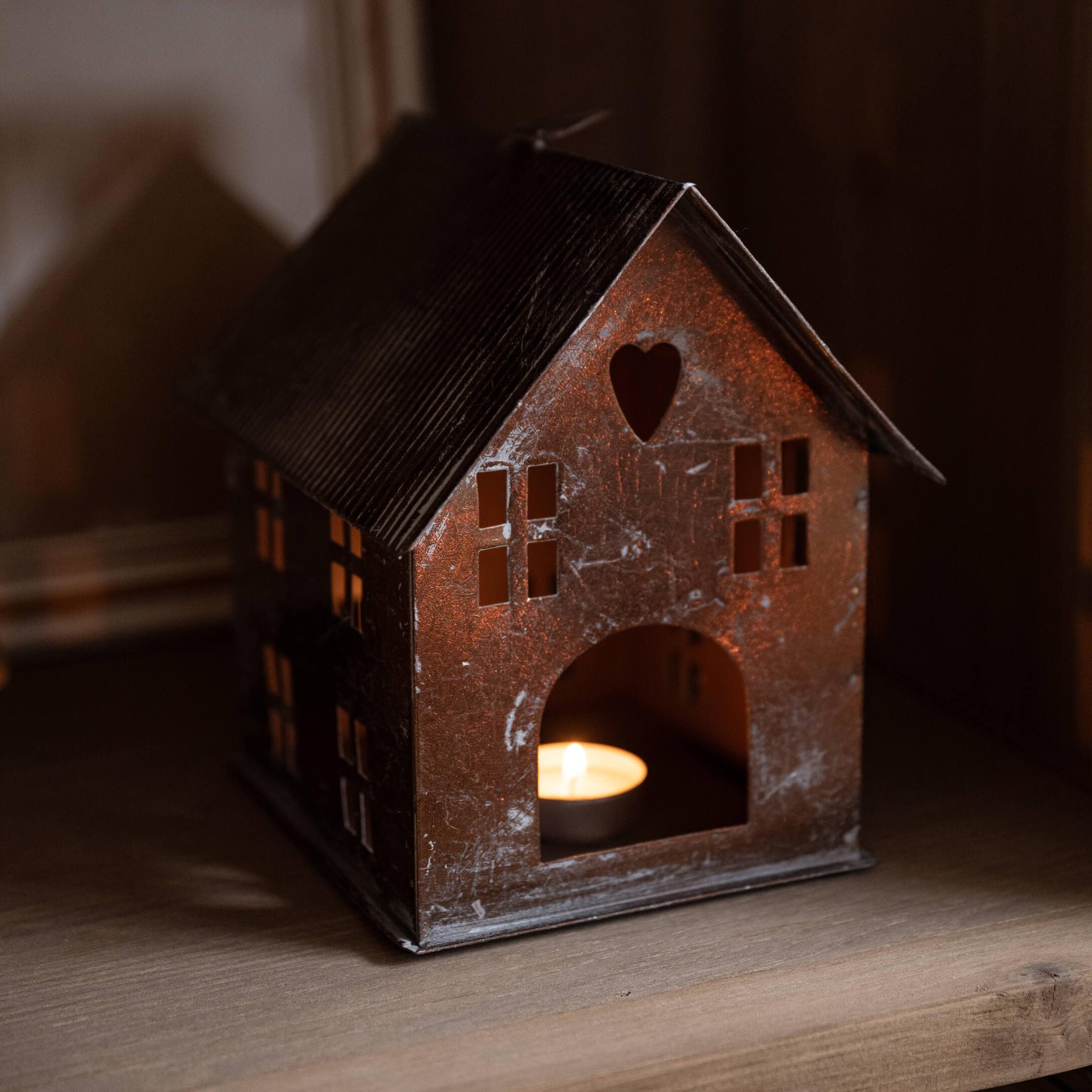 Metall-Laterne in Hausform mit Herz-Ausschnitt und brennender Kerze innen auf Holzregal