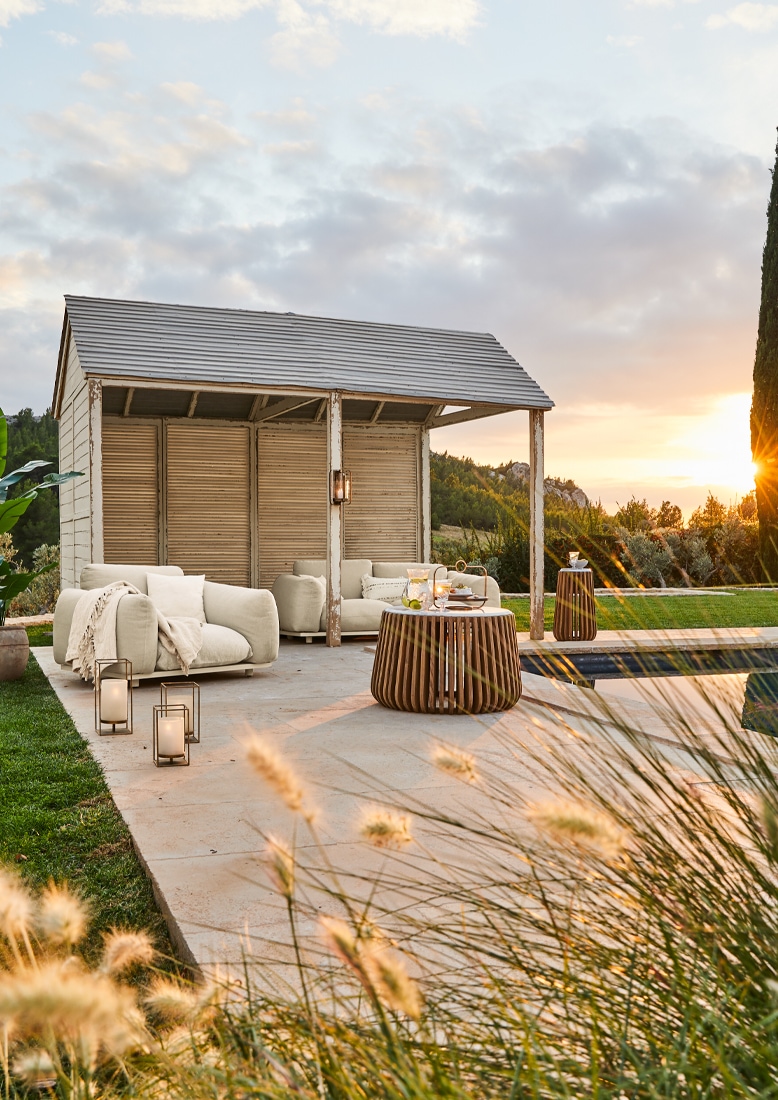 Gemütlicher weißer Gartenpavillon mit beigen Möbeln und Holztisch auf Terrasse bei Sonnenuntergang