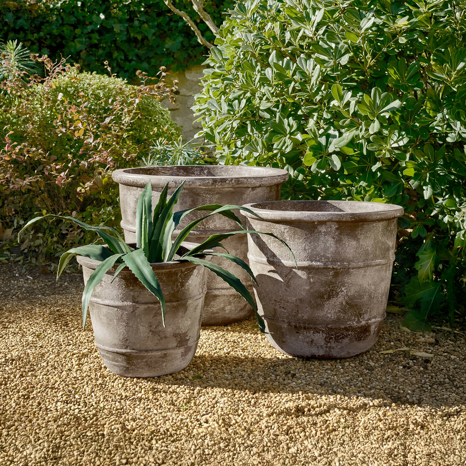 Drei große graue Steintöpfe auf Kiesboden im Garten mit grünen Pflanzen im Hintergrund