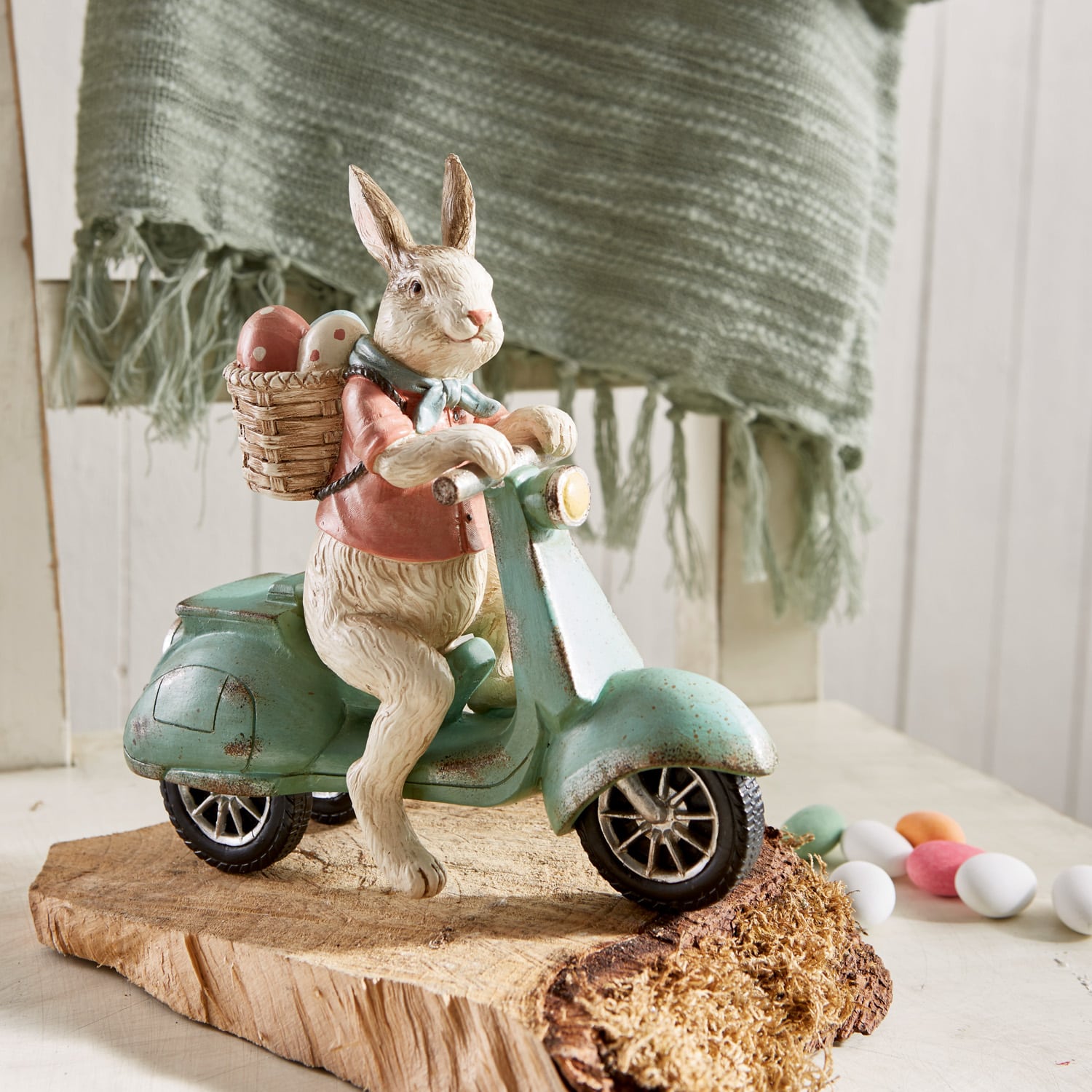 Hasenfigur auf grünem Roller mit Korb voller bunter Ostereier auf Holzsockel