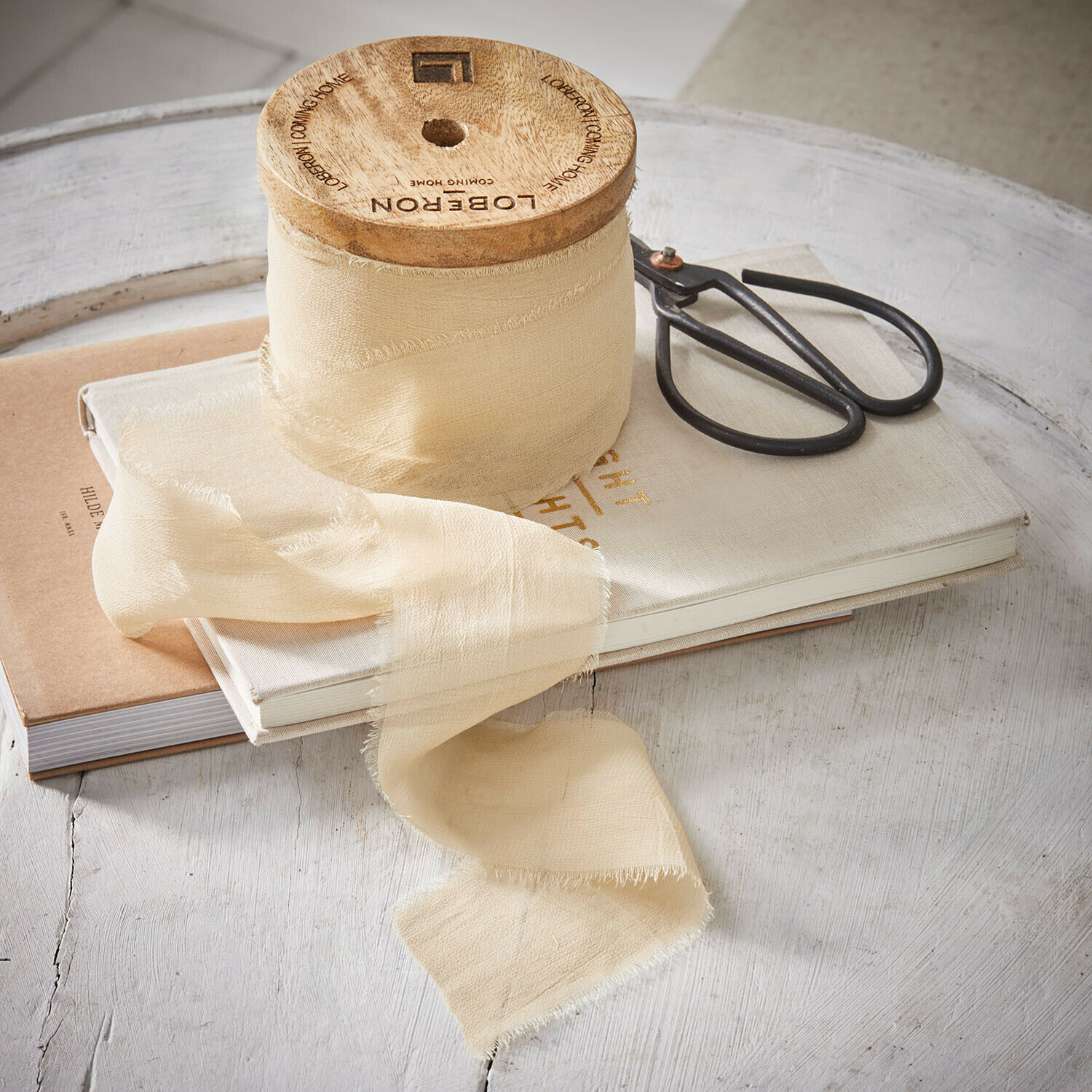 Beige Stoffband aus Naturmaterial auf Holzspule neben Schere und Büchern auf weißem Tisch