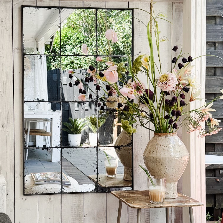 Wandspiegel mit geteilten Paneelen, der Blumen in Keramikvase und Kaffee auf rustikalem Holztisch reflektiert