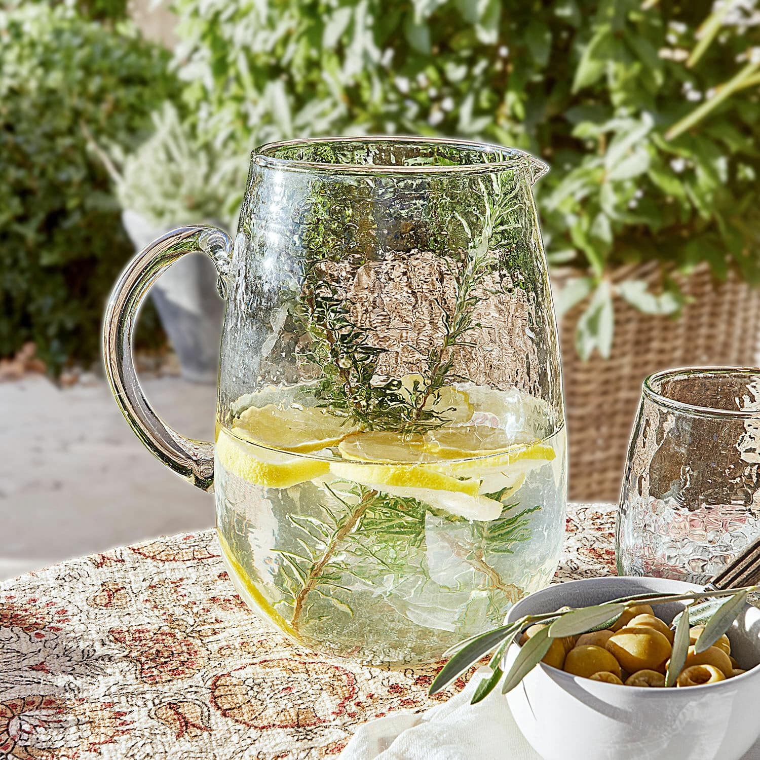 Krug Rezay Glaskrug mit Wasser, Zitrone und Kräutern auf gemustertem Tischtuch, erfrischendes Sommergetränk