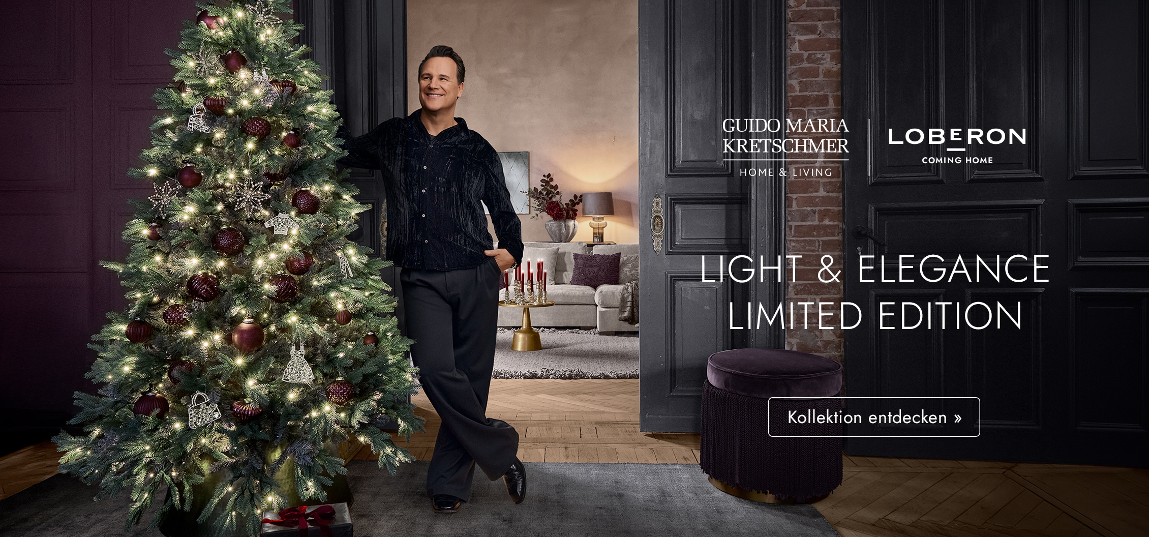 Mann lehnt an beleuchtetem Weihnachtsbaum mit bordeauxroten und silbernen Kugeln in elegantem Wohnzimmer