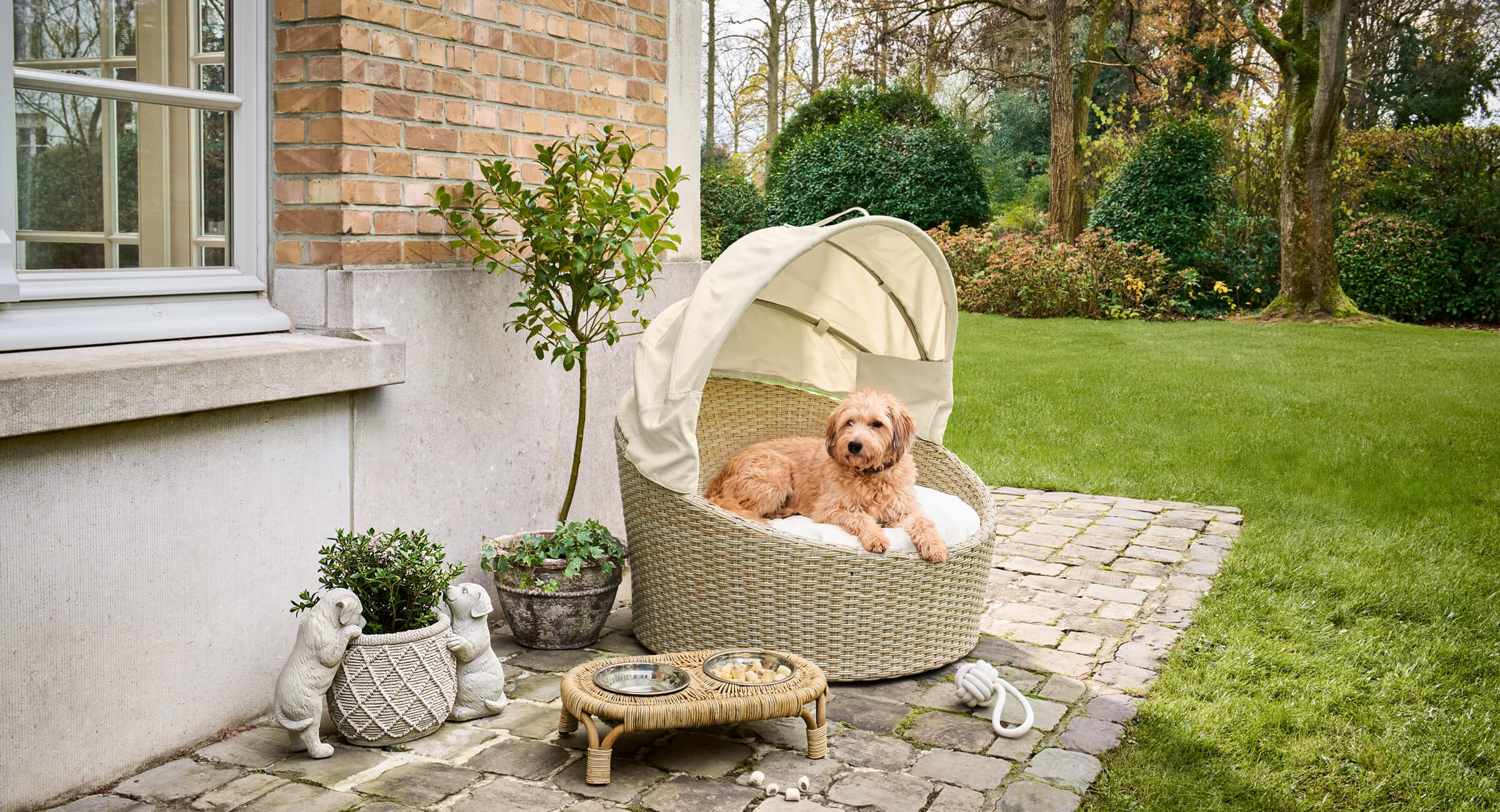 Beige geflochtener Hundekorb mit Dach auf Steinterrasse, gemütlicher Rückzugsort im Garten