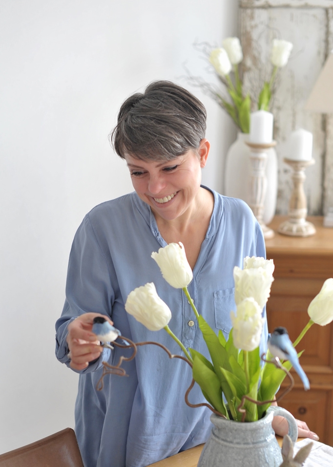 Frau in blauem Hemd mit weißen Tulpen und dekorativen Vögeln in grauem Krug auf dem Tisch
