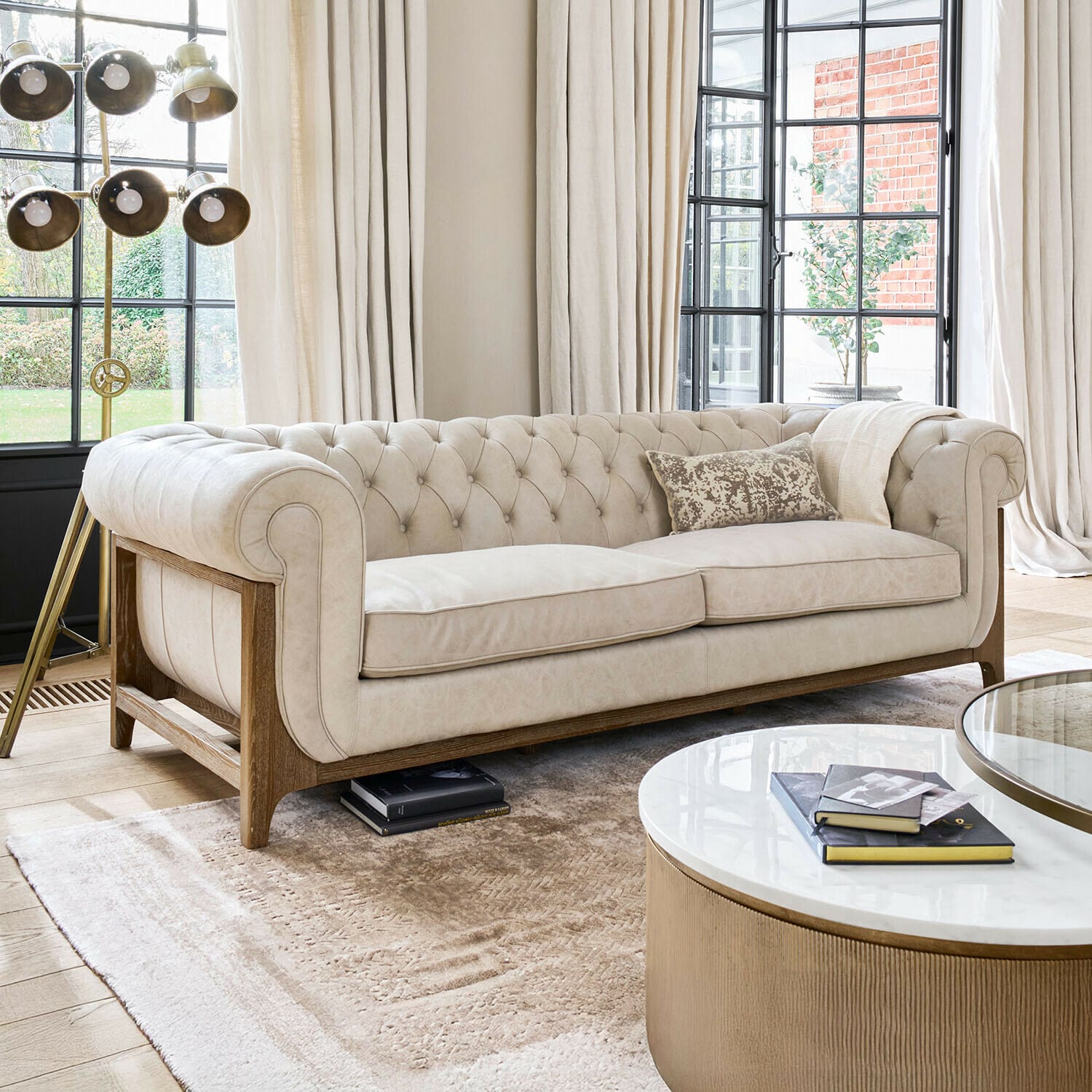 Sofa Swansboro Beige gepolstertes Chesterfield-Sofa mit Holzrahmen in hellem Wohnzimmer mit großen Fenstern und Stehlampe