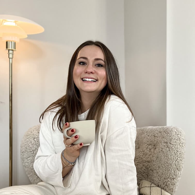 Lächende Frau im weißen Pullover hält eine beige Tasse, sitzt auf weichem Sessel in hellem Raum