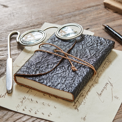 Schwarzes Notizbuch aus strukturiertem Leder mit Schnurverschluss auf Holztisch mit Brille und Feder