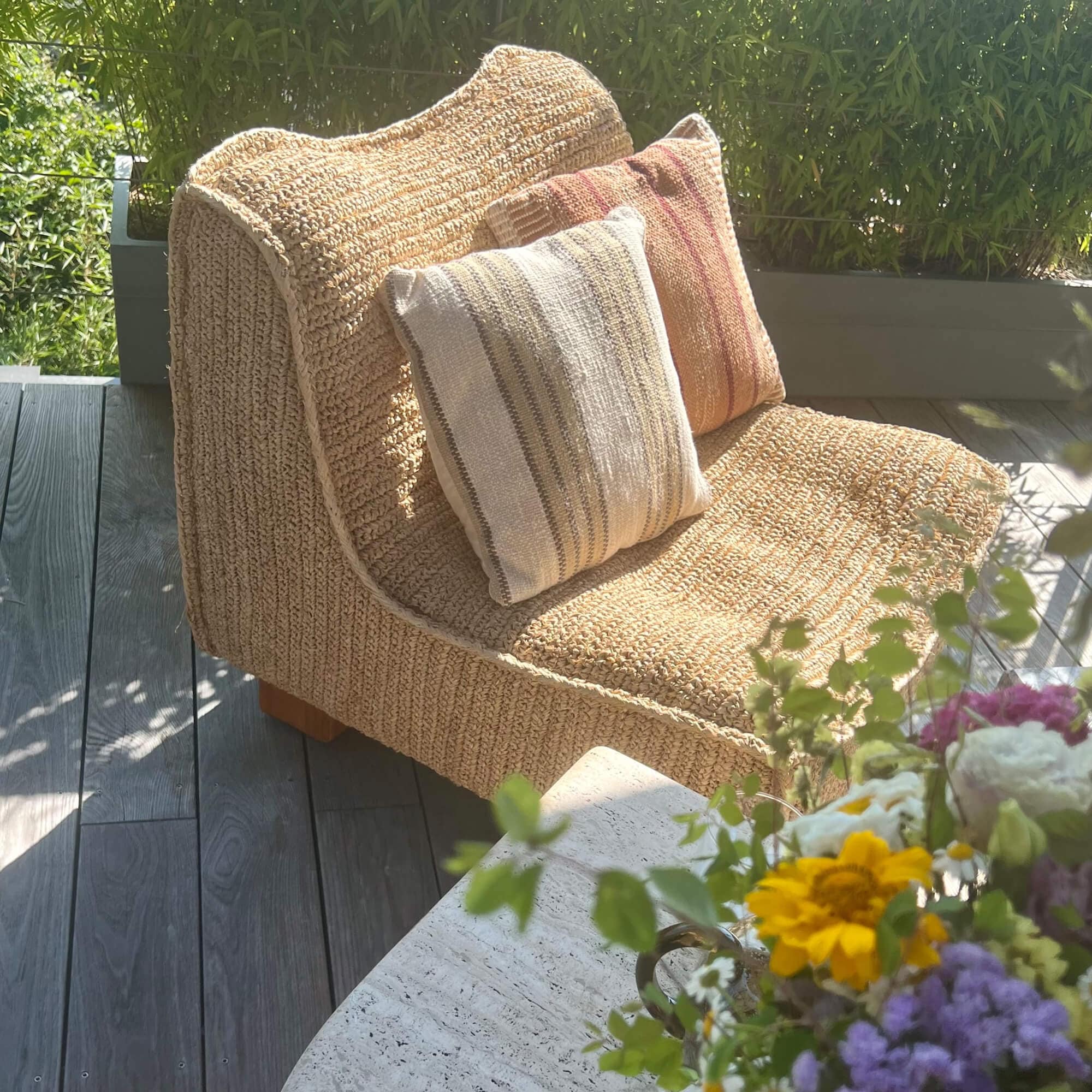 Beiger Sessel aus geflochtenem Material mit zwei dekorativen Kissen auf Terrasse mit Blumen