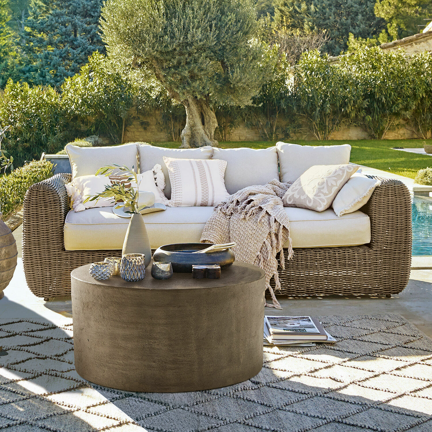 Sofa Bogdanne Beiges Rattan-Gartensofa mit Kissen und rundem Betontisch auf gemustertem Teppich im Garten