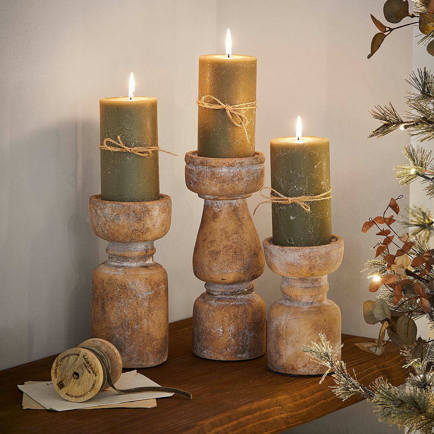 Drei rustikale Holzkerzenhalter mit grünen Kerzen und Juteschnur auf dekorativem Holzregal