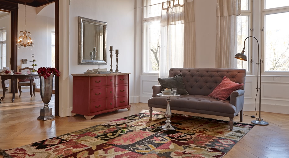 Wohnzimmer im French Chic mit grauem, gepolstertem Sofa, rotem Holzkommode und buntem Teppich
