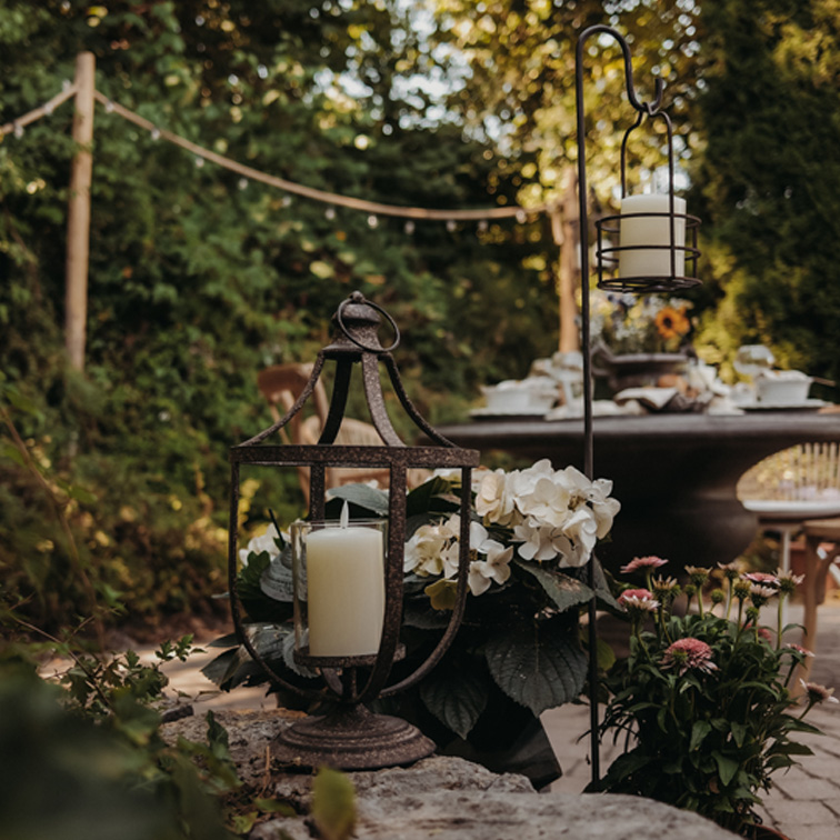Metalllaterne mit weißer Kerze auf Steinmauer im Garten mit Blumen und Tisch im Hintergrund