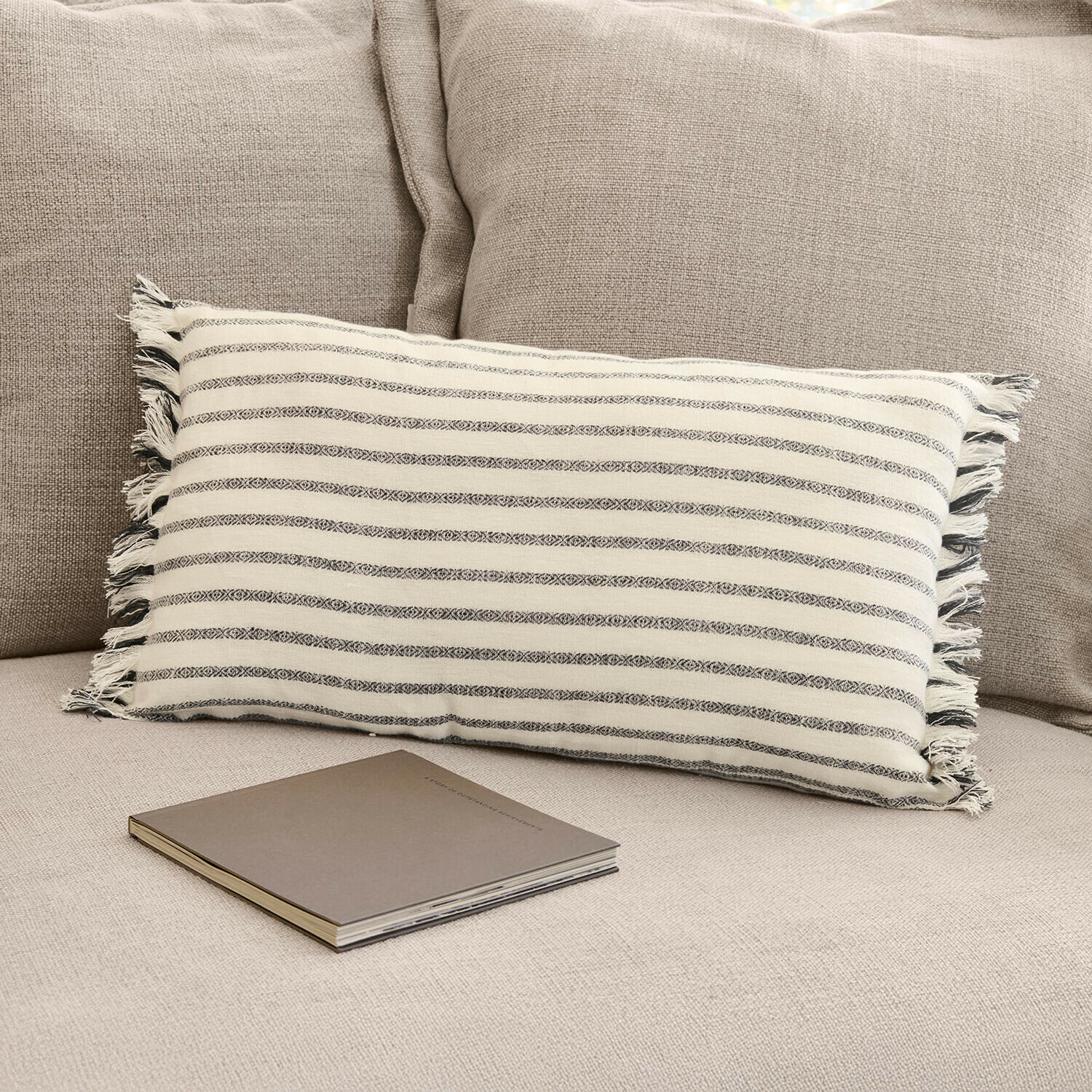 Kissenhülle Ulderina Rechteckiges Baumwollkissen mit creme-grauen Streifen auf beigem Sofa mit Buch